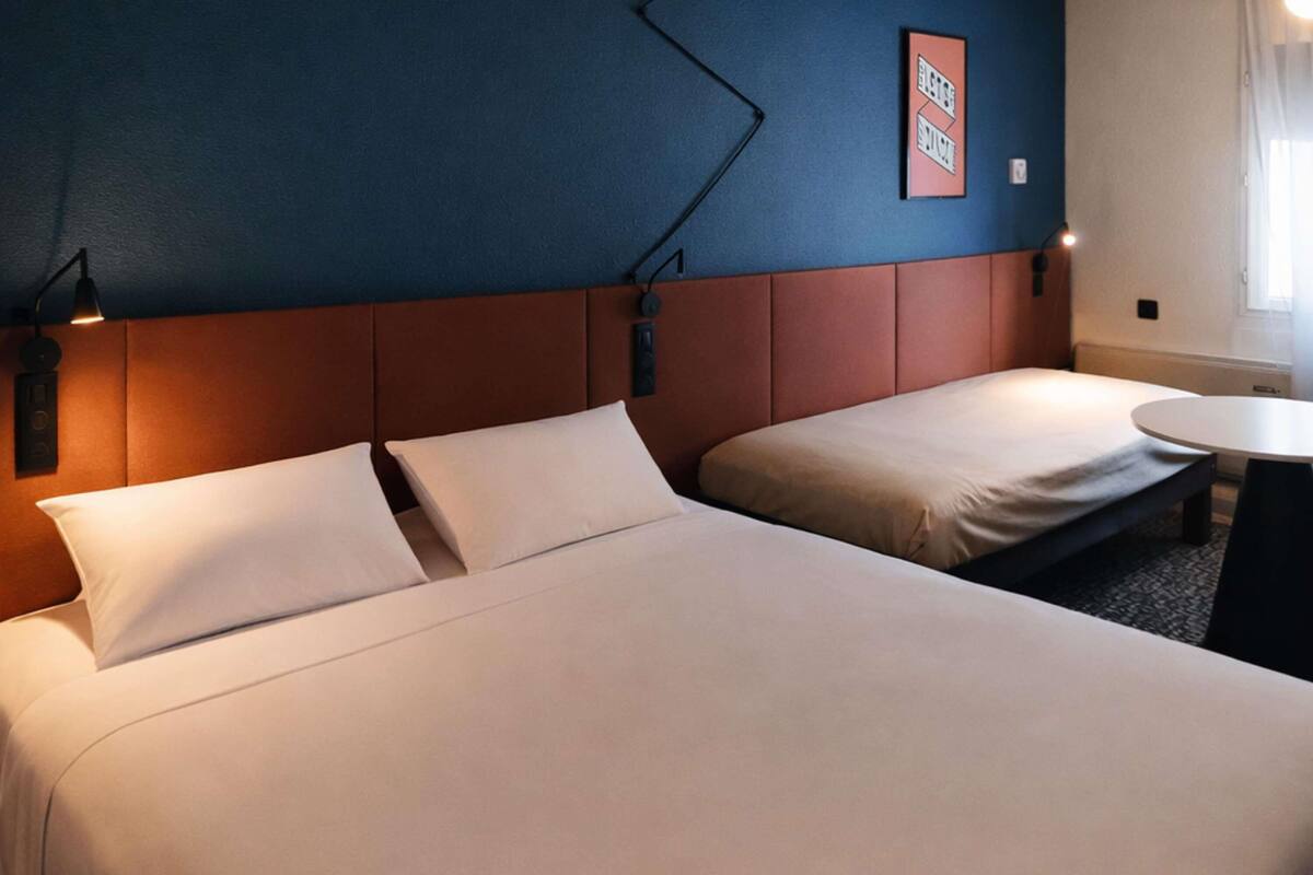 A hotel room with two beds, a round table, and a wall-mounted light fixture.