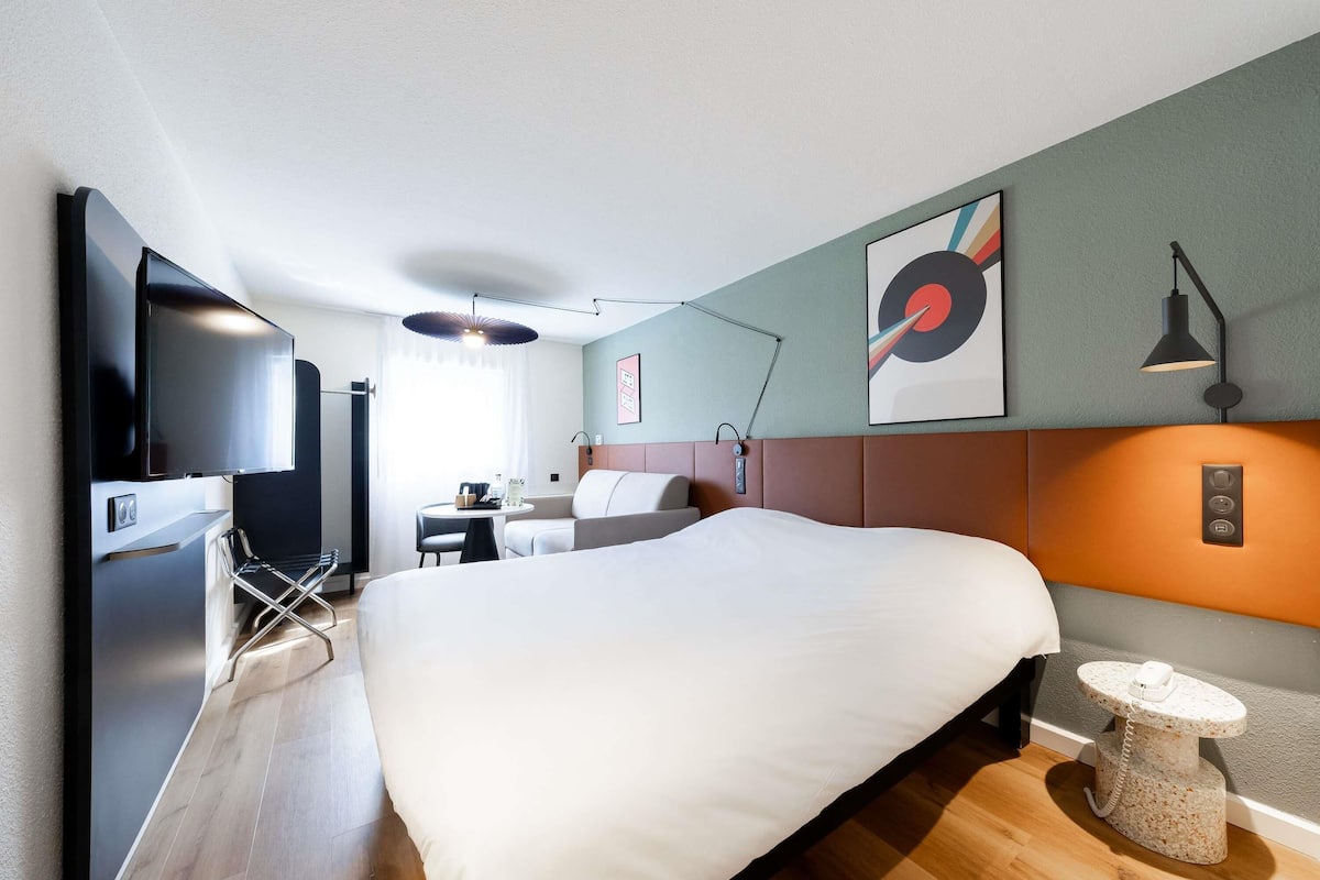 A modern hotel room with a large bed, a flat-screen TV, a desk with a chair, and a small round table with a phone.