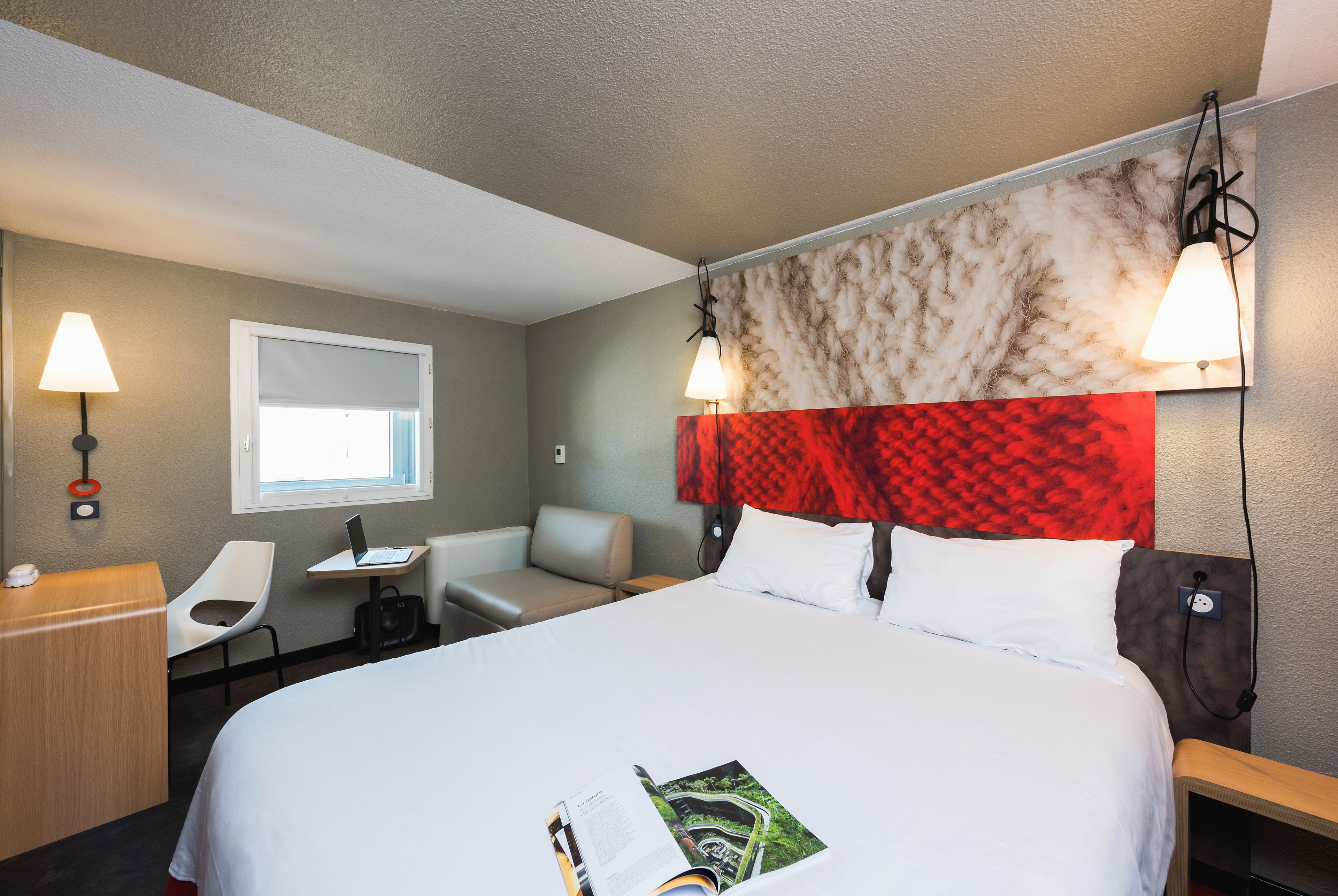 Quarto de hotel com uma cama grande, uma escrivaninha com um laptop, uma cadeira e uma janela.