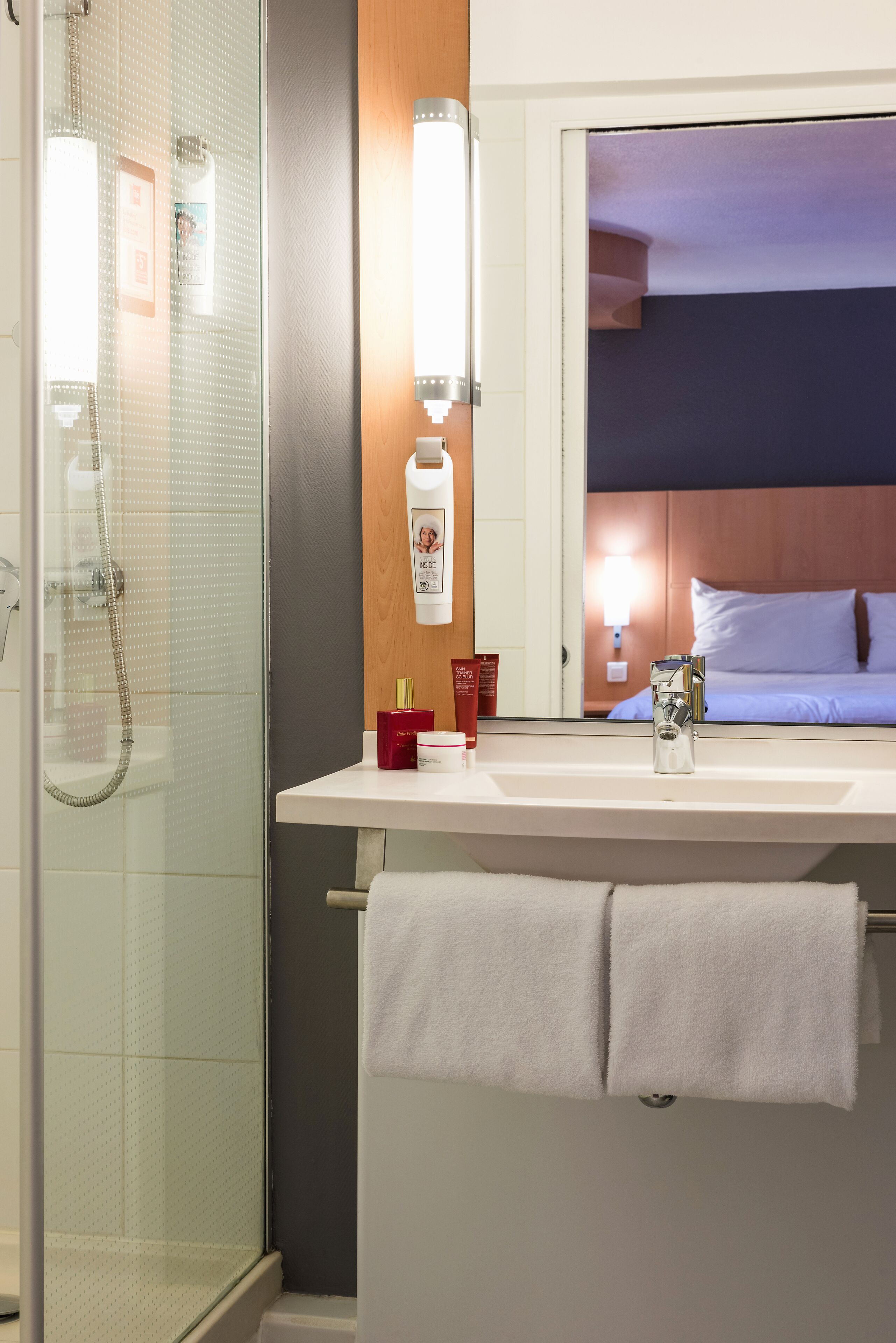 Ein Hotelbad mit einer gläsernen Duschkabine, einem Waschbecken mit Spiegel darüber und einem Handtuchhalter mit zwei Handtüchern.