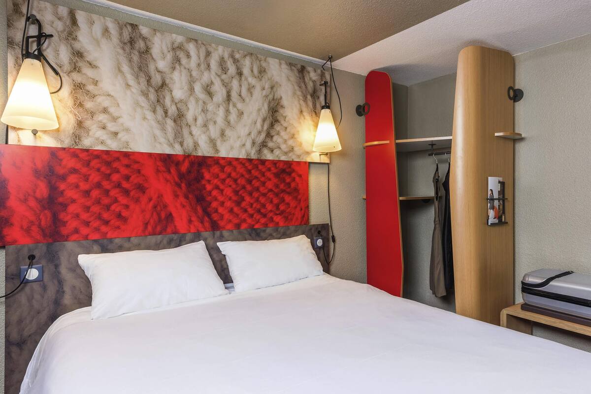 A hotel room with a bed, two wall-mounted lamps, a red and beige headboard, a wooden wardrobe, and a shower area.