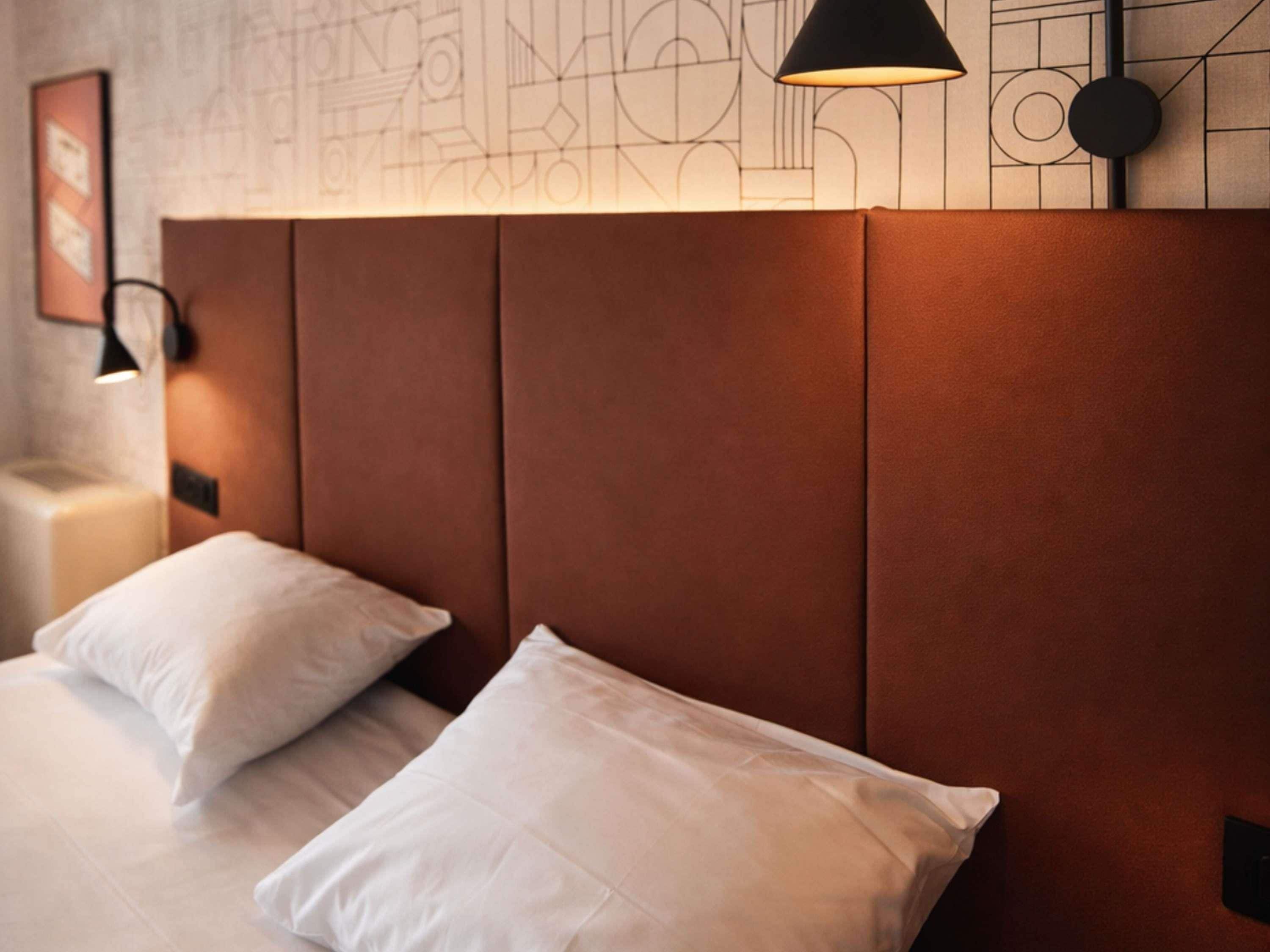 A hotel room with a bed, pillows, a nightstand, a lamp, and a wall with geometric patterns.