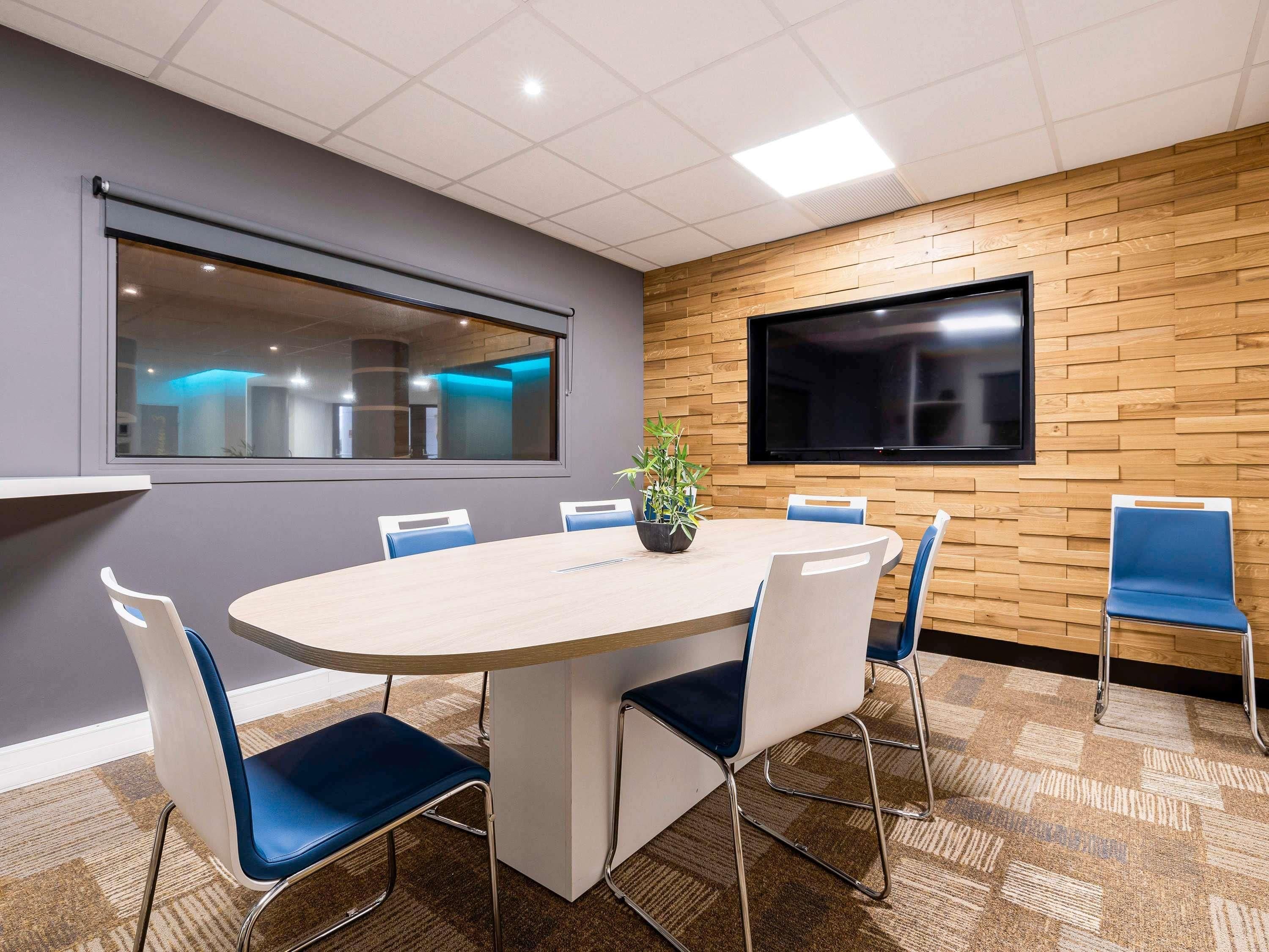 A conference room with a round table, blue chairs, a potted plant, a large TV, and a window with a view of the interior.