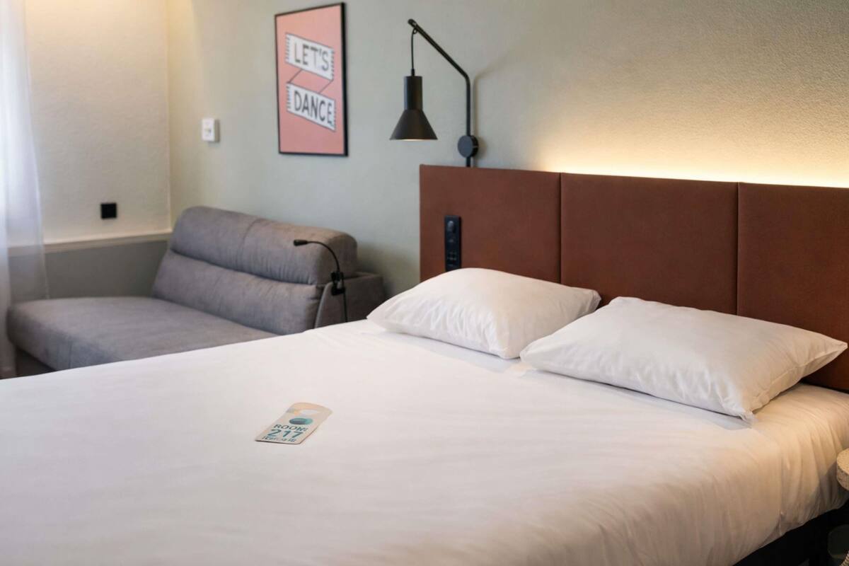 A hotel room with a bed, a grey armchair, a wall-mounted lamp, and a sign that says 'LET'S DANCE'.