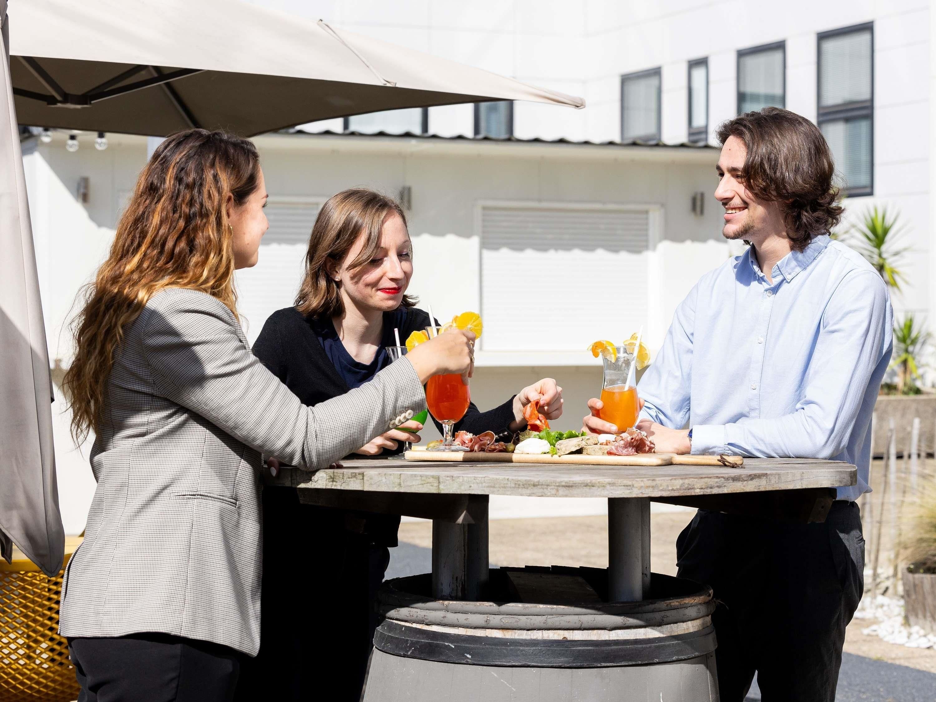 Tres personas sentadas en una mesa al aire libre con comida y bebidas, bajo un toldo.