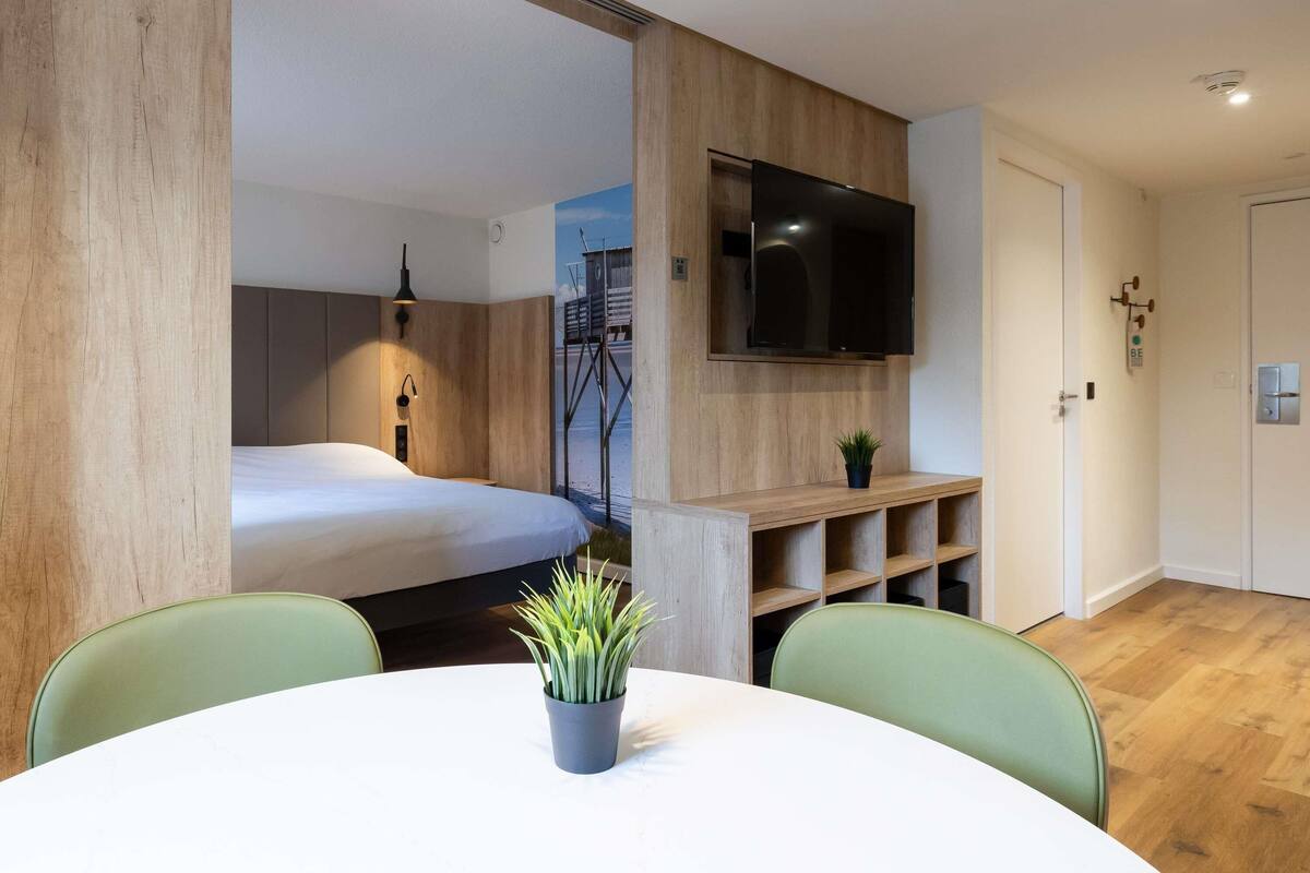 A modern room with a bed, a TV mounted on the wall, a small plant on a shelf, and a round table with chairs.
