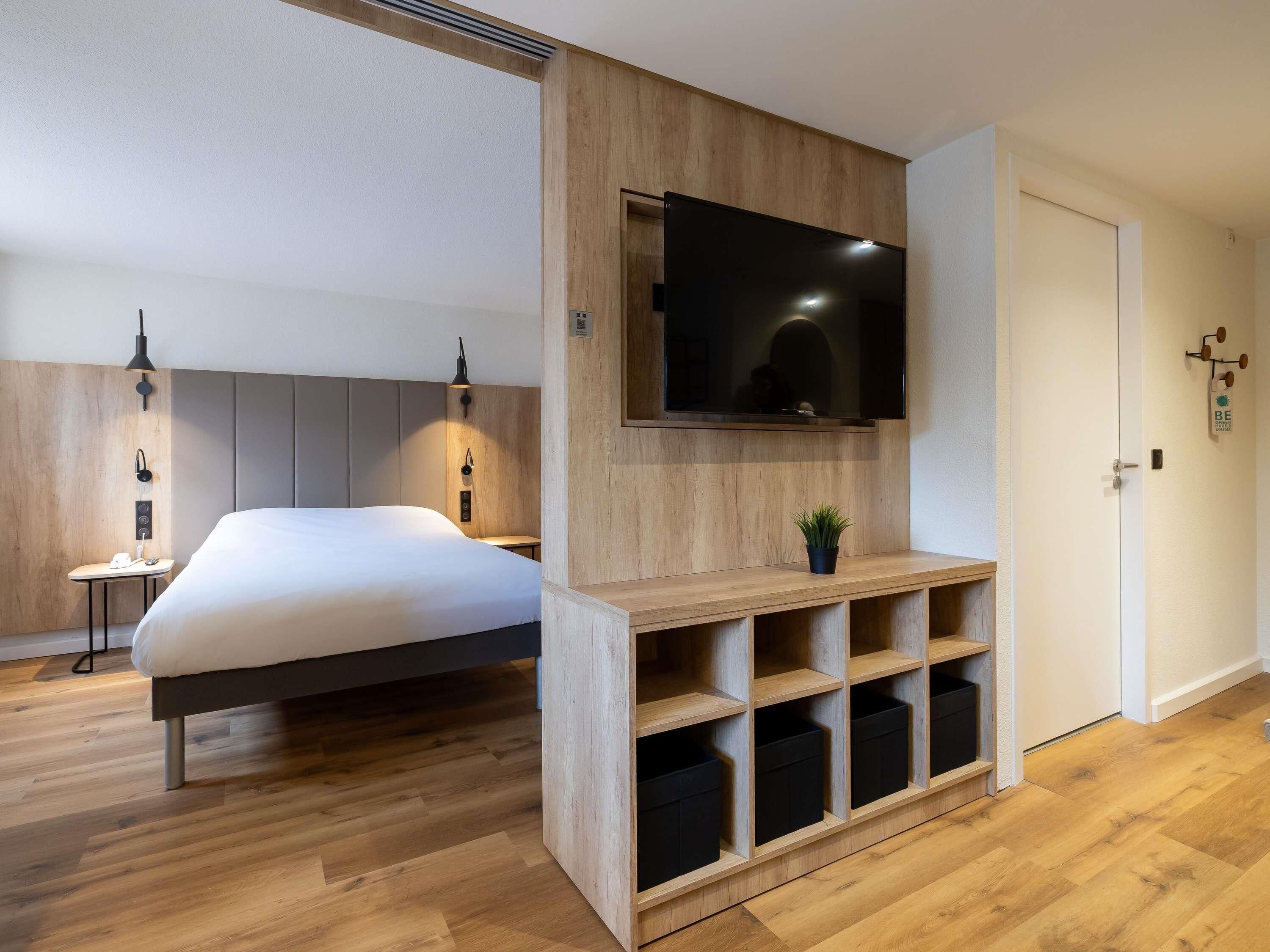 Ein modernes Hotelzimmer mit einem Bett, einem an der Wand befestigten Fernseher, eingebauten Regalen und einem Nachttisch mit Lampe.