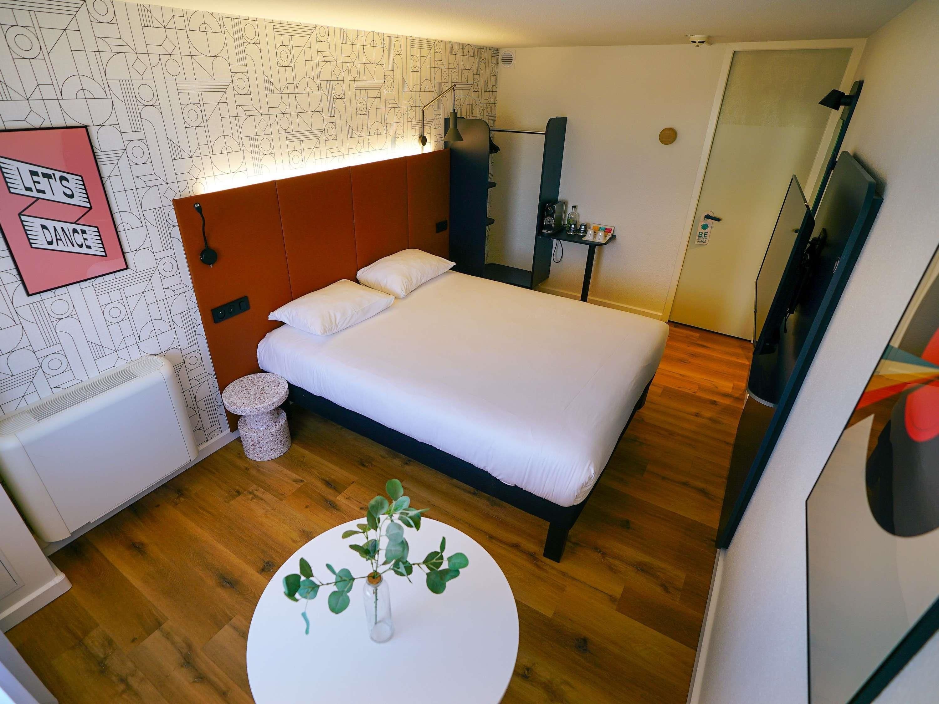 A modern bedroom with a bed, a bedside table, a mirror, and a wall with geometric patterns.