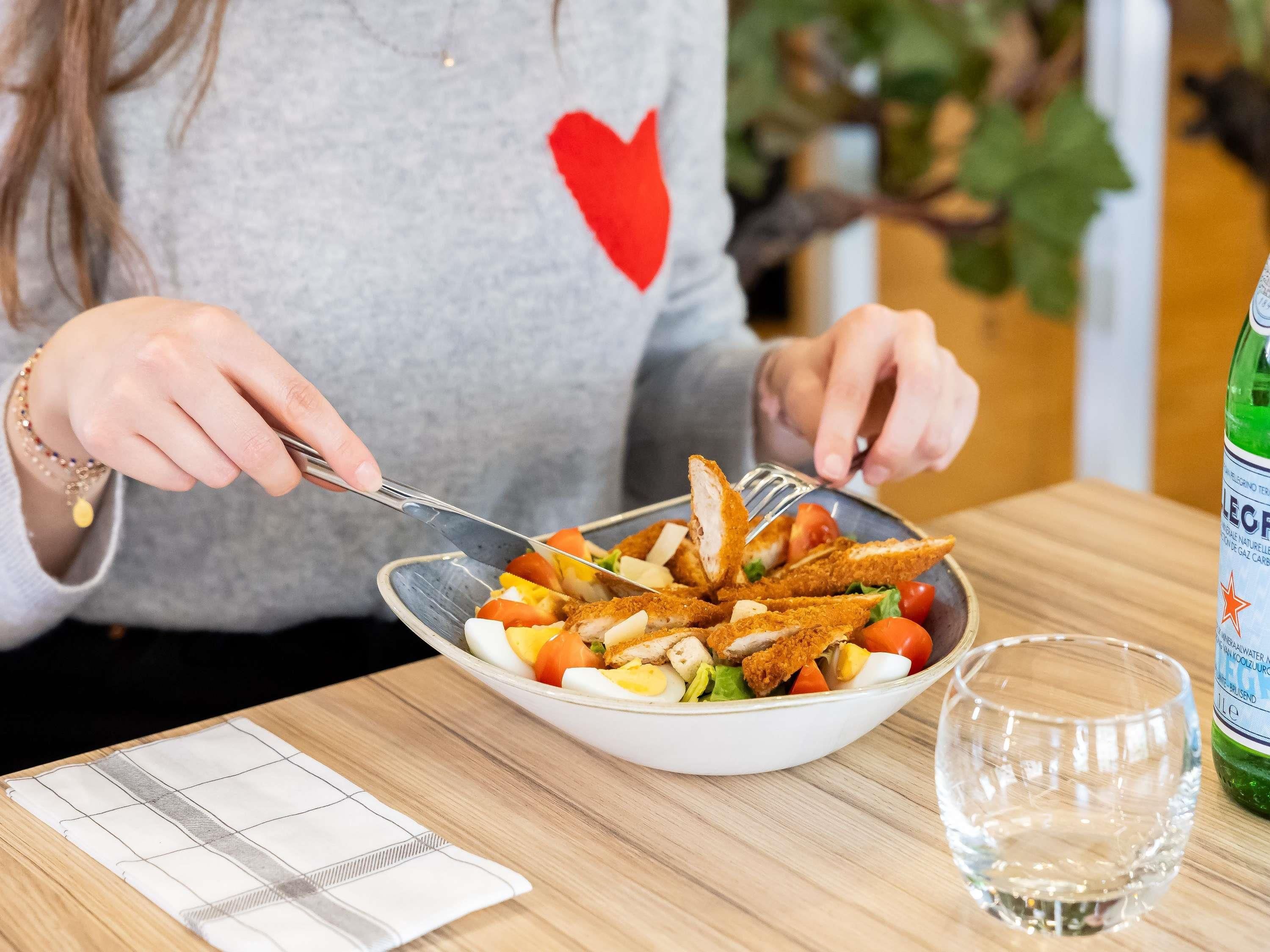 Une personne est assise à table et prend un repas avec une fourchette et un couteau, un verre d’eau et une bouteille d’eau à portée de main.