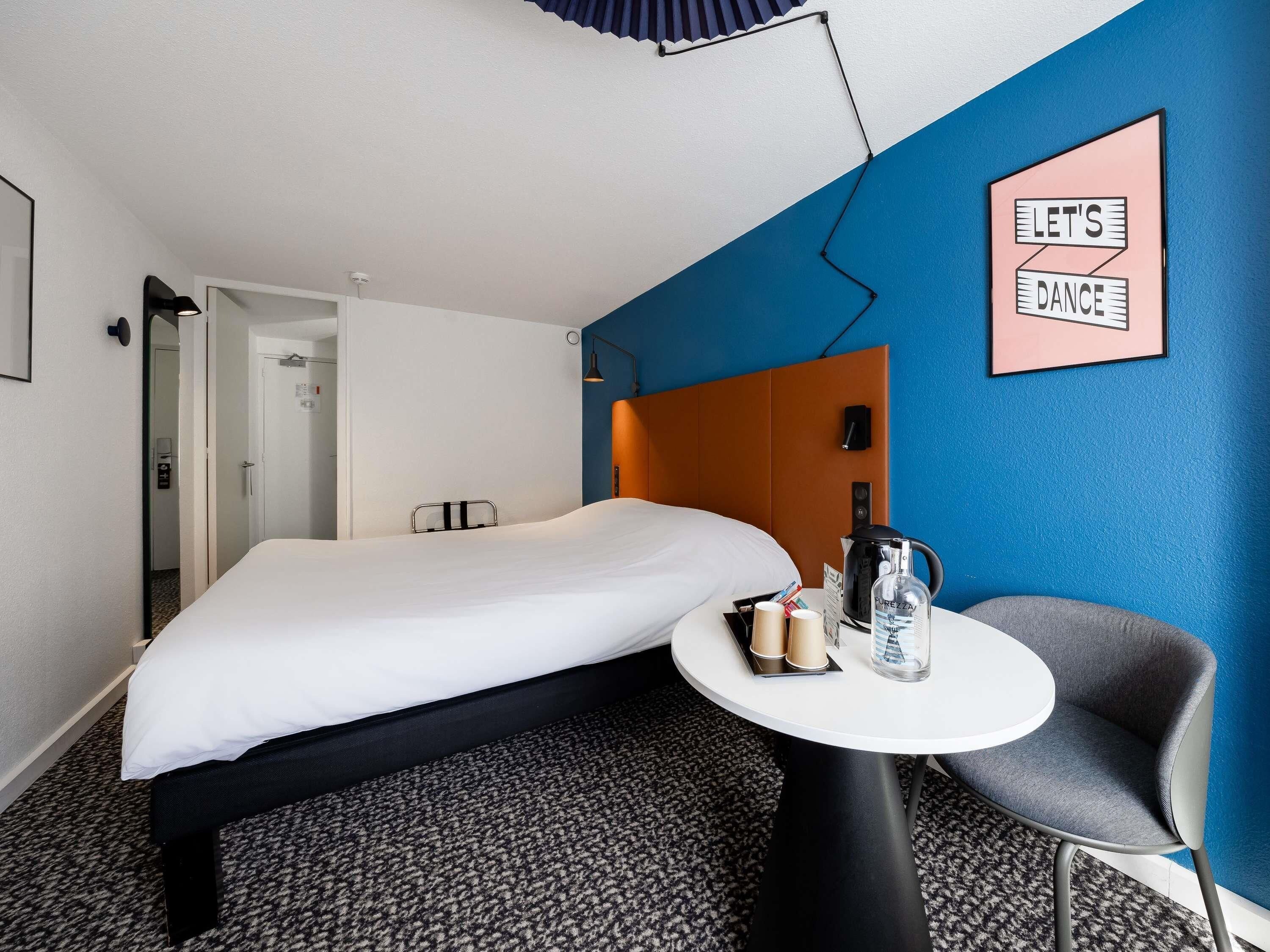 Una camera d'albergo con un letto, un tavolo con un bollitore, una sedia e un cartello che recita 'LET'S DANCE'.