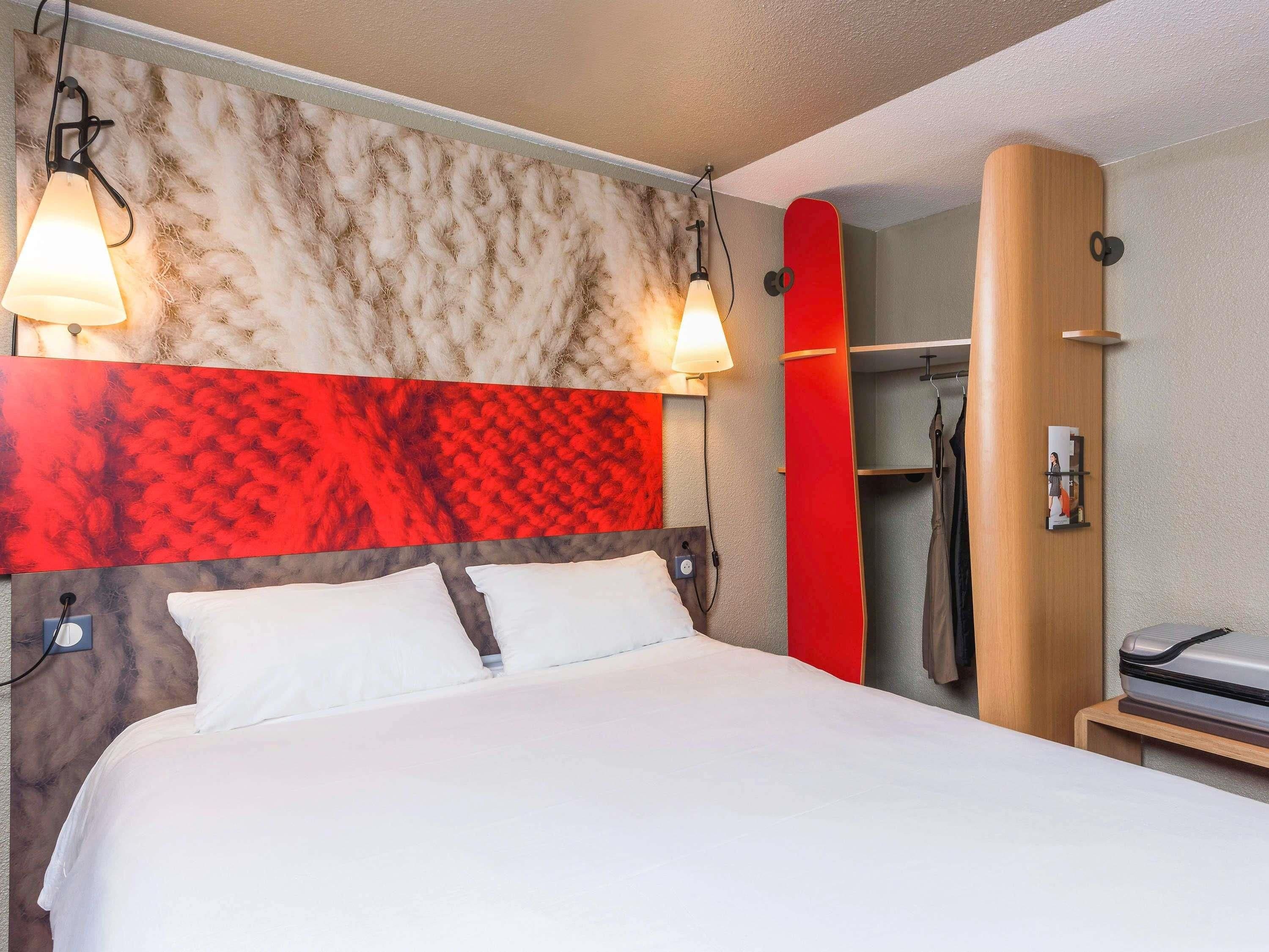 A hotel room with a bed, a headboard with a red and beige pattern, a wooden wardrobe, and a shower area.