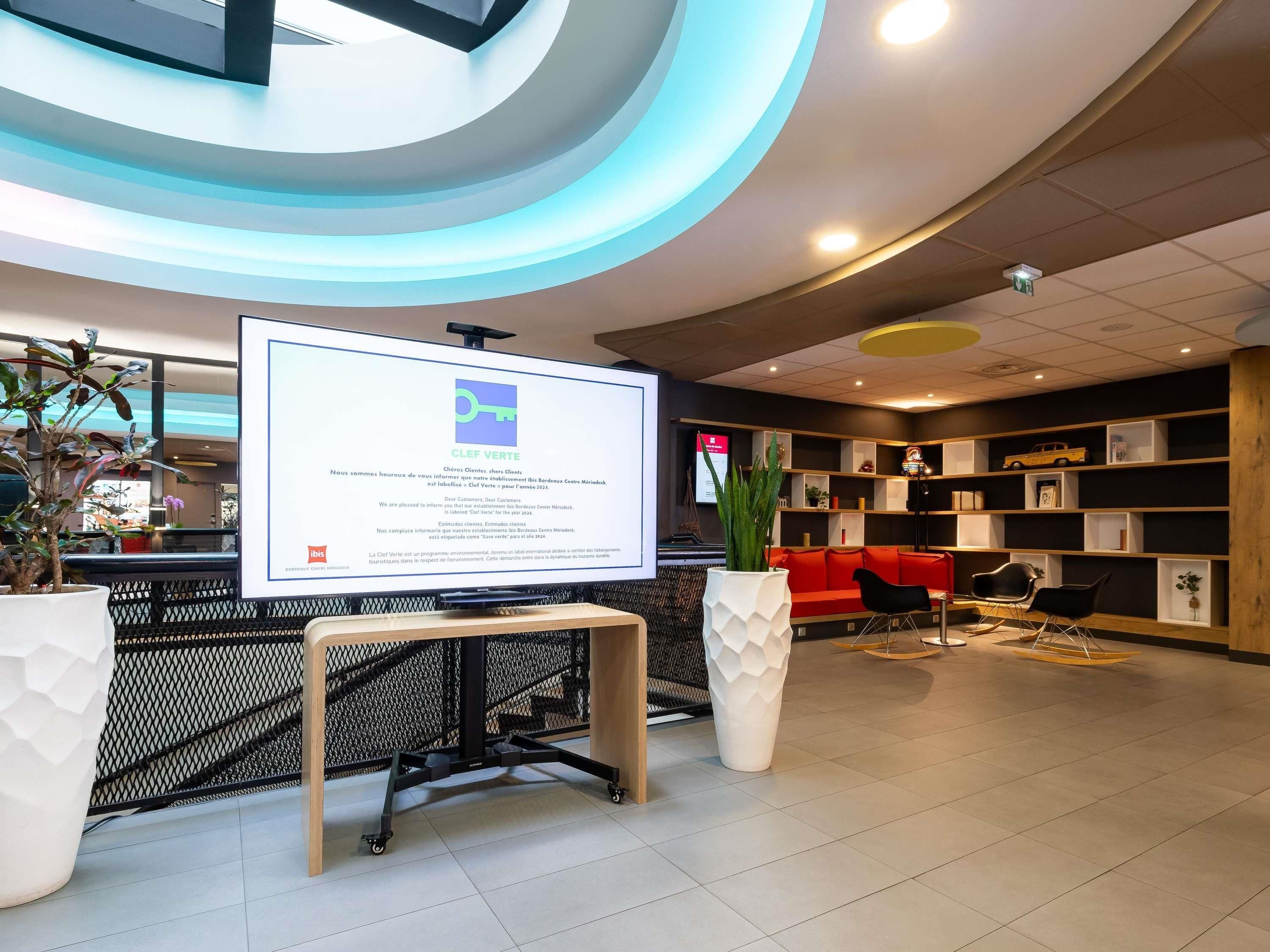 Un espace d’accueil moderne doté d’un écran numérique affichant des informations, d’un coin salon avec un canapé rouge et d’une bibliothèque.