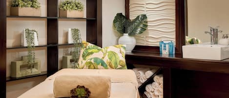 Un cuarto de spa con una camilla de masaje, estantes con plantas, un espejo grande y un lavamanos con artículos de tocador.
