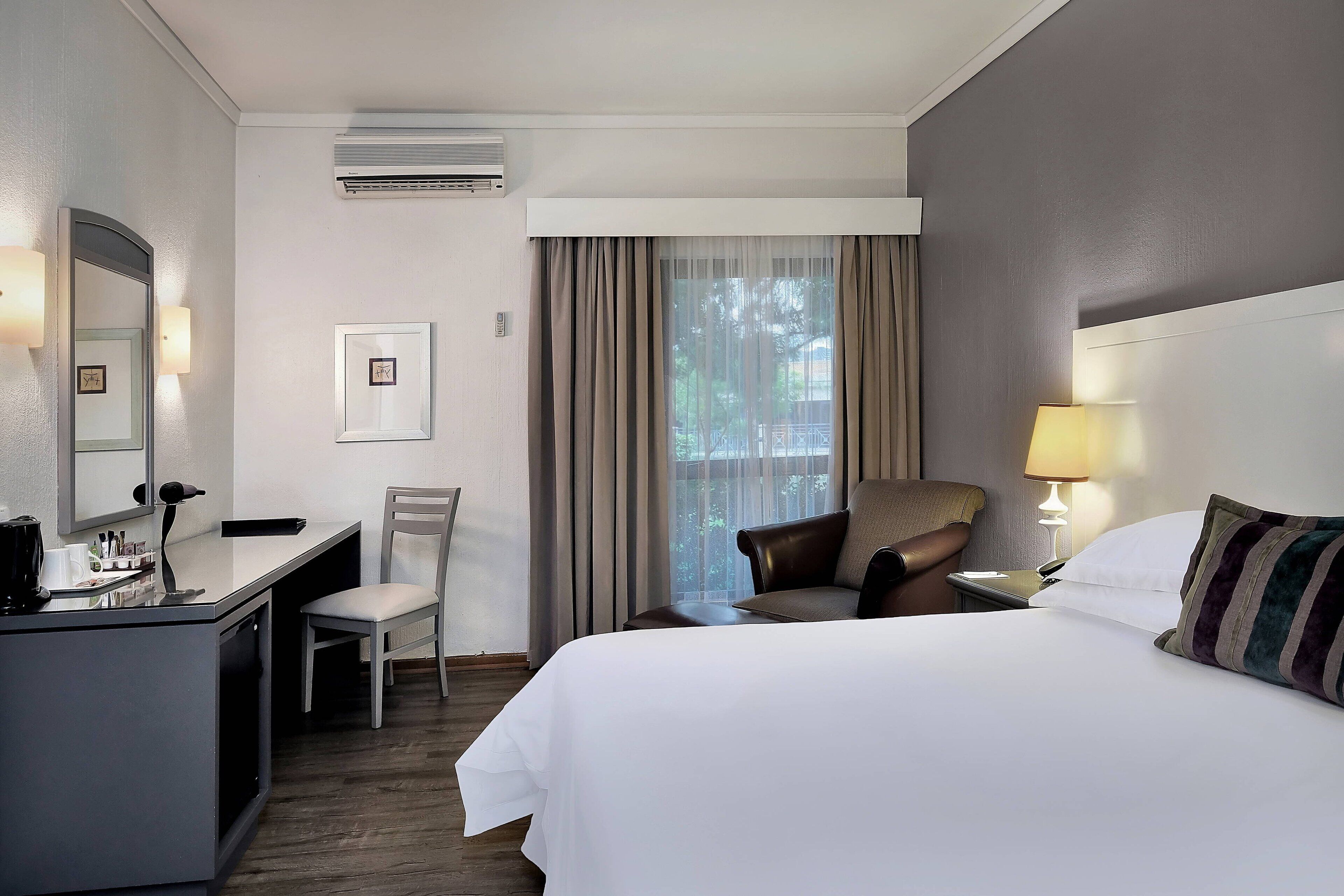A hotel room with a bed, a desk, a chair, a lamp, and a window with curtains.