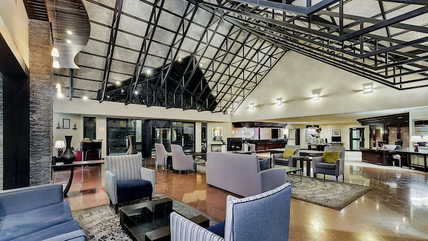 A spacious lobby with modern furniture, a high ceiling with a geometric design, and a visible kitchen area. - Protea Hotel by Marriott Midrand (Midrand)