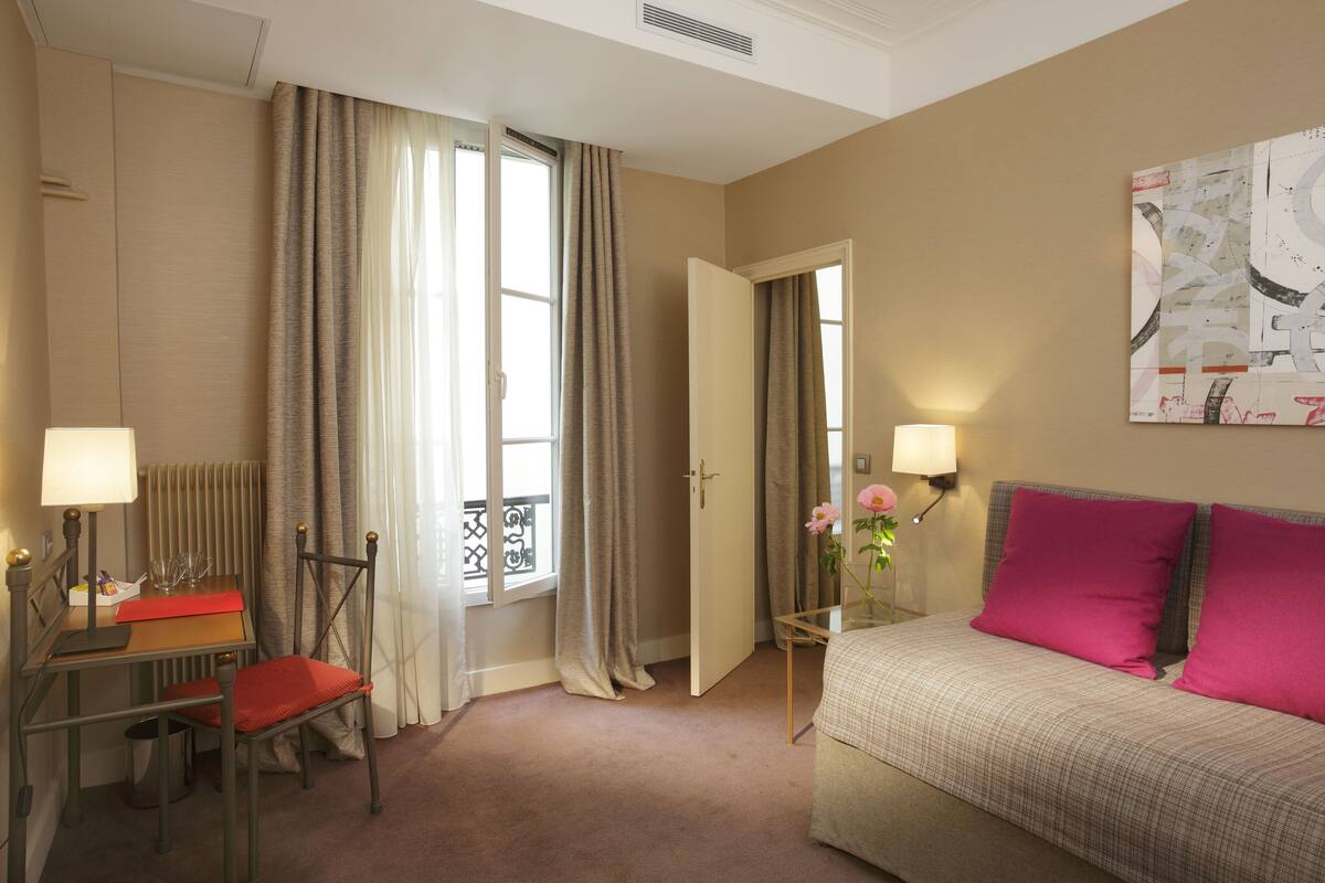 Een hotelkamer met een bed, een bureau met stoel, een lamp, een raam met gordijnen en een schilderij aan de muur.