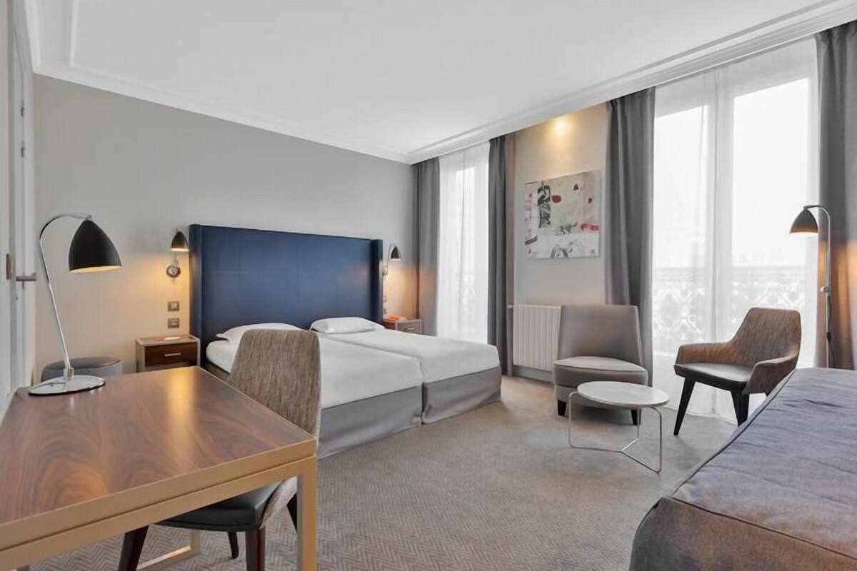 Een moderne hotelkamer met een groot bed, een bureau, een stoel en een bank. Er is een raam met gordijnen en een schilderij aan de muur.