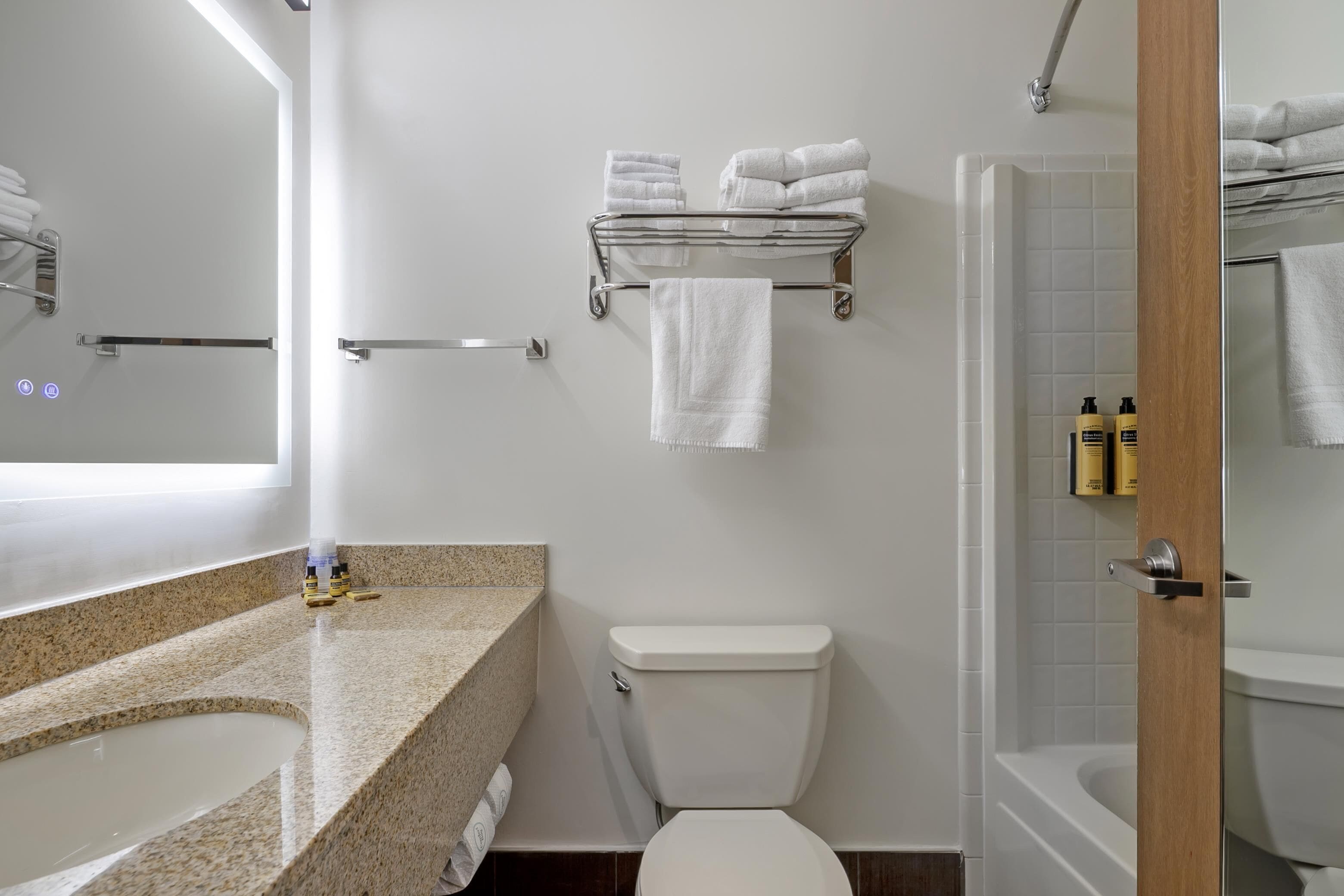Un baño con encimera de granito, inodoro, repisa de vidrio con toallas y zona de ducha.