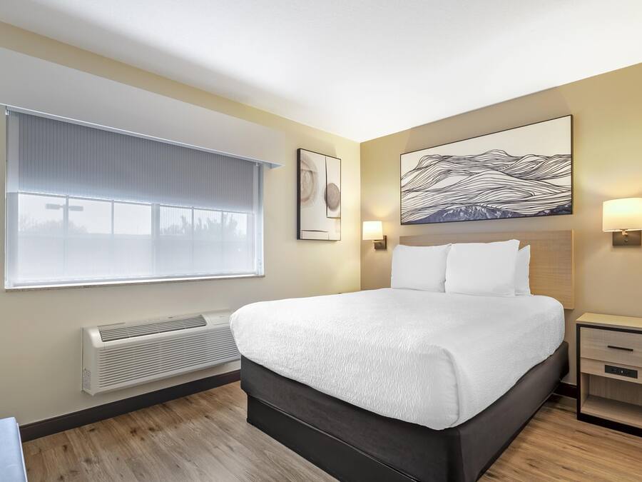 A hotel room with a large bed, a window with blinds, a wall-mounted heater, a bedside table with a lamp, and a painting on the wall.