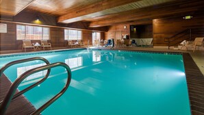 An indoor swimming pool with wooden walls and a tiled floor. There are lounge chairs and a small table near the pool.