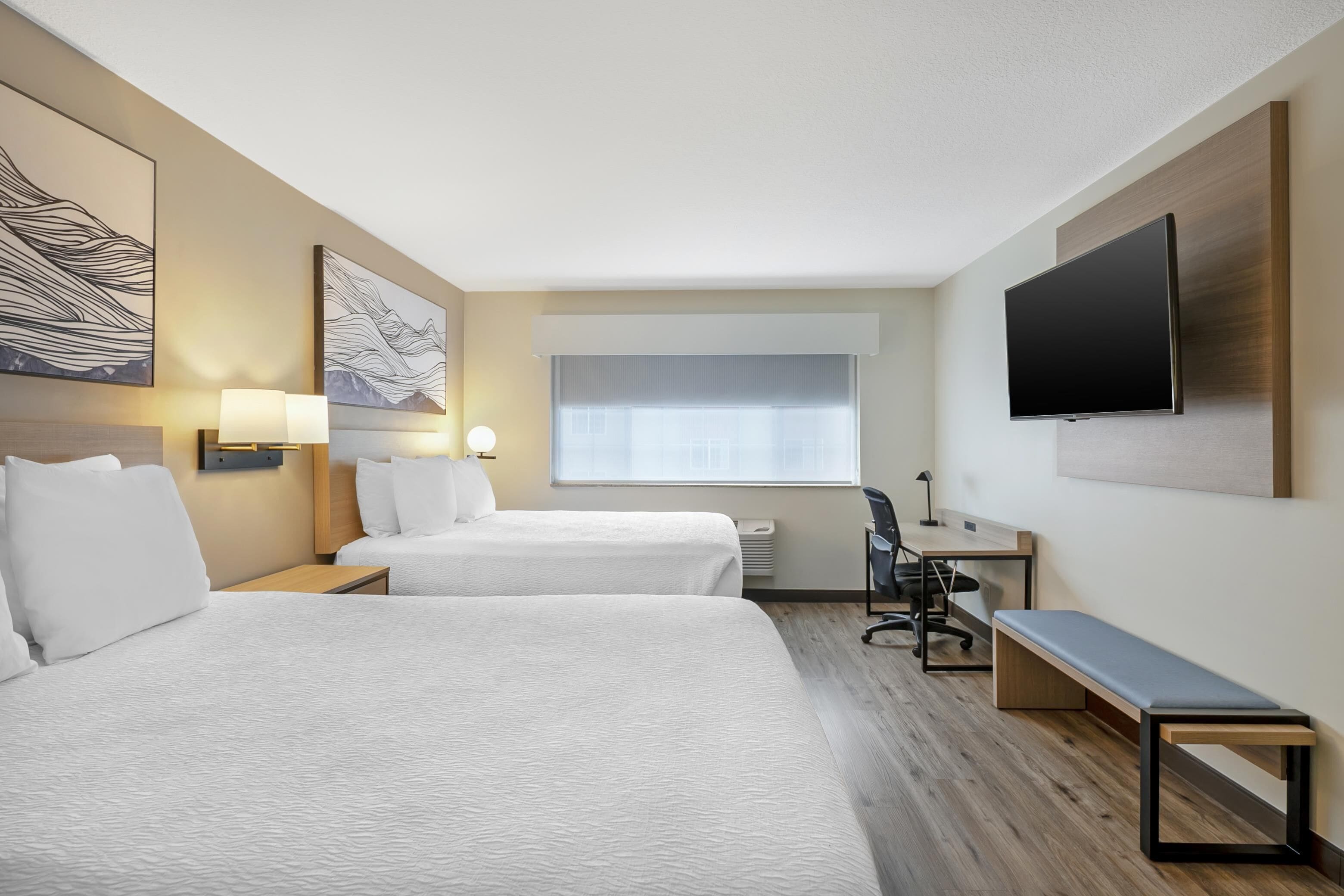 A hotel room with two beds, a desk, a chair, a TV, and artwork on the walls.