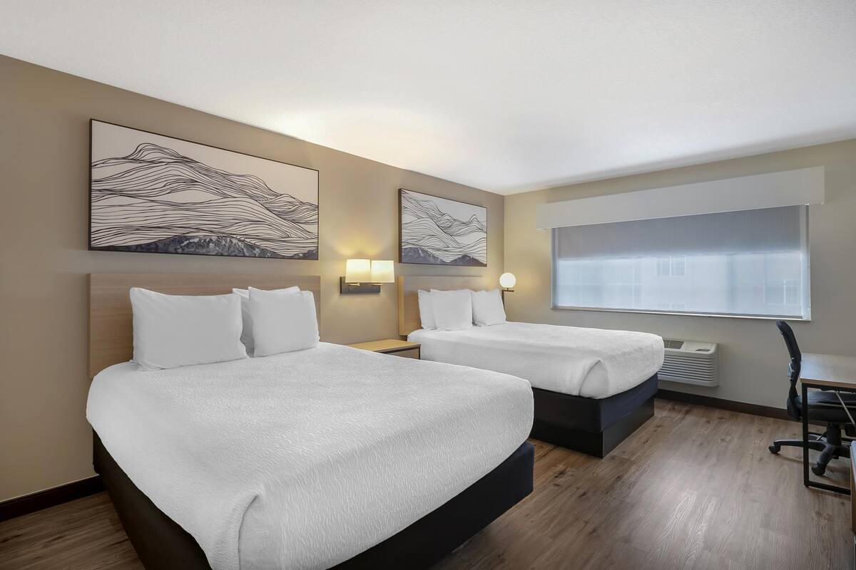 A hotel room with two beds, wall-mounted artwork, bedside lamps, a window with blinds, and a desk with a chair.