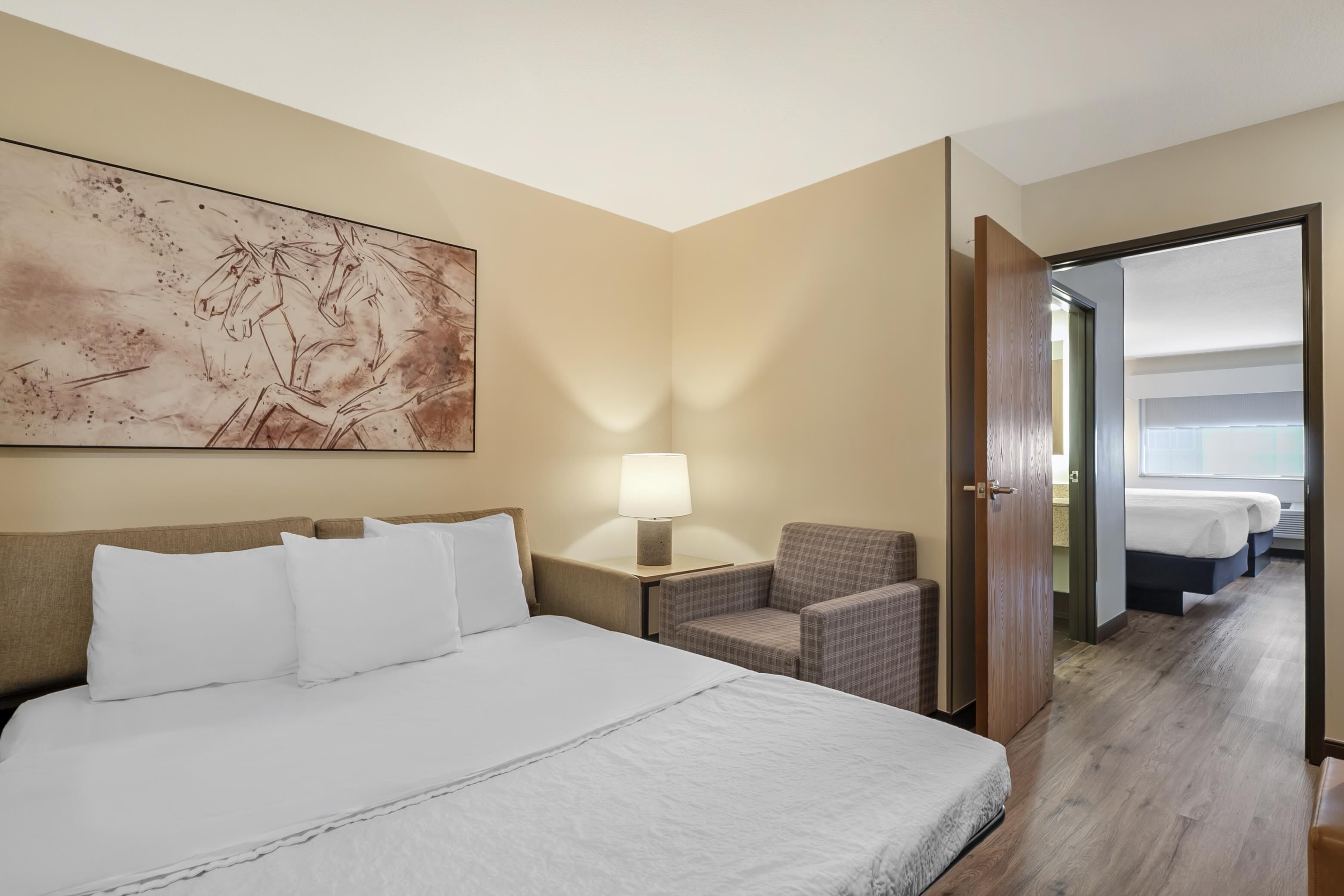 A hotel room with a large bed, a chair, a nightstand with a lamp, and a painting on the wall.