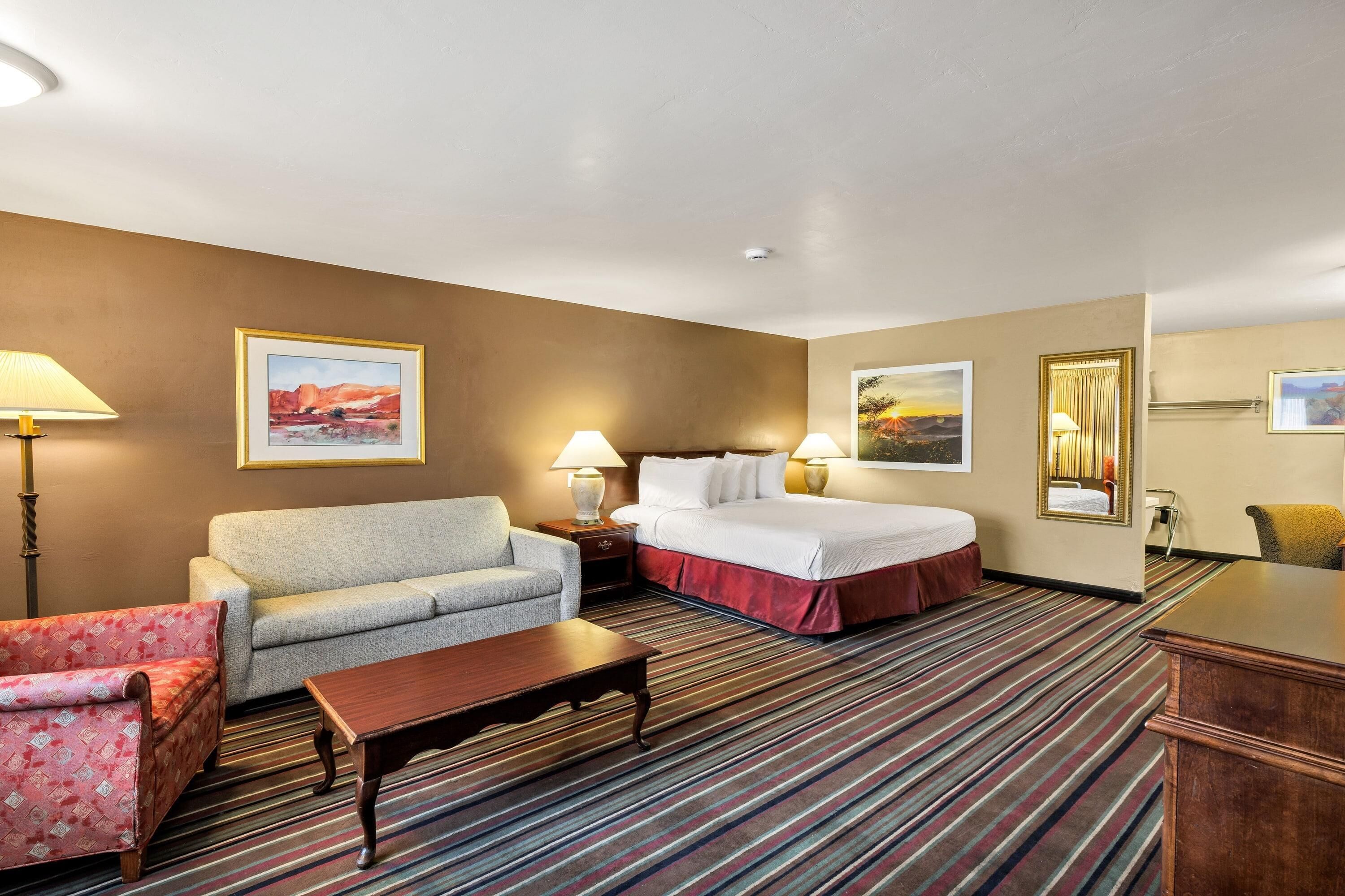 Quarto de hotel com uma cama, um sofá, uma mesa pequena e uma escrivaninha. Há duas luminárias nas mesas de cabeceira e um espelho na parede.
