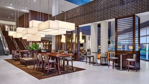 A spacious lobby with a high ceiling, modern lighting fixtures, a central seating area with tables and chairs, and a staircase leading to upper levels.