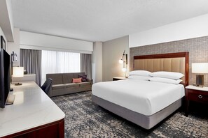 A hotel room with a large bed, a sofa, a desk with a TV, and a window with curtains. - Sheraton Indianapolis Hotel at Keystone Crossing (Indianapolis)