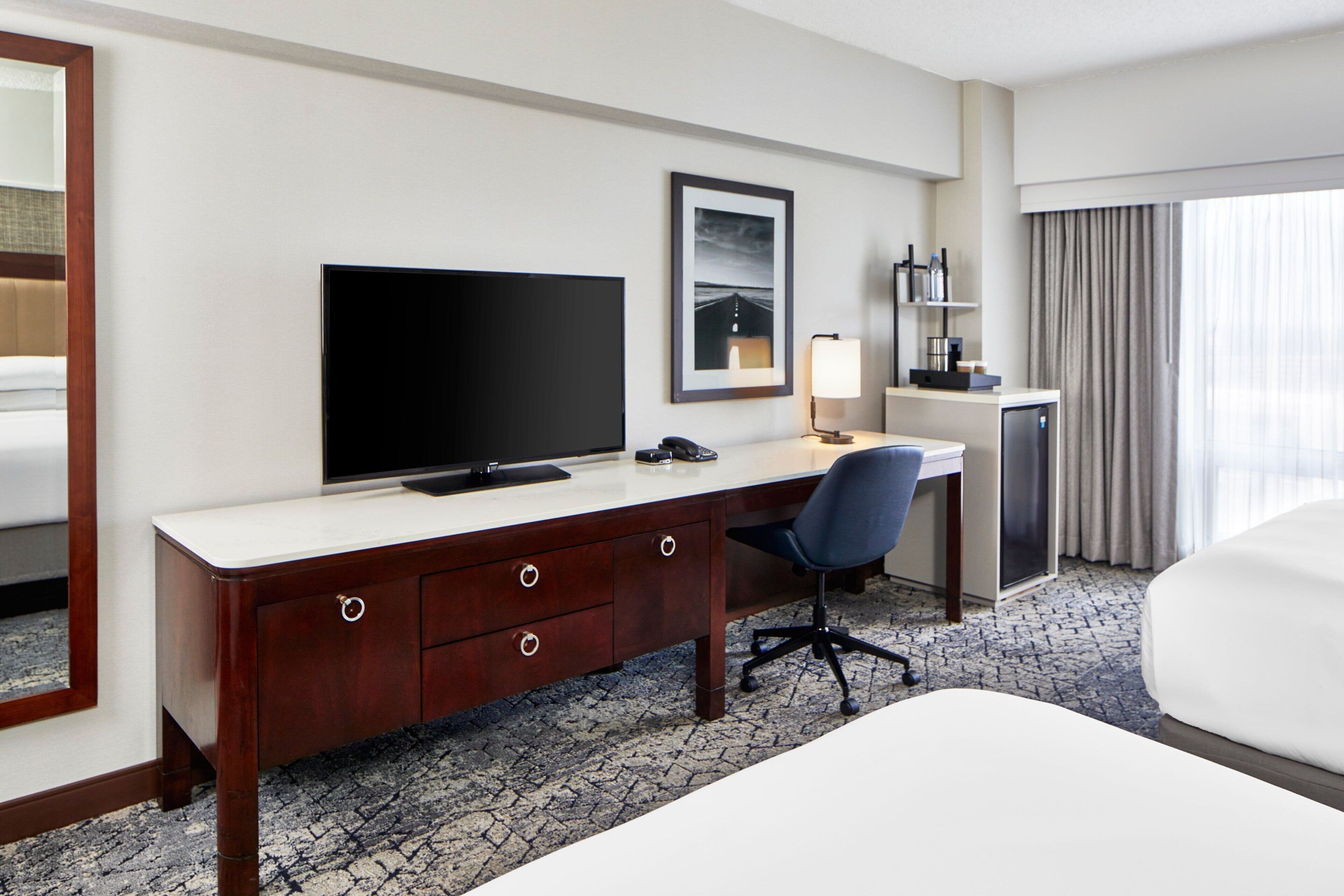 A hotel room with a large bed, a desk with a computer, a TV, and a small kitchenette.