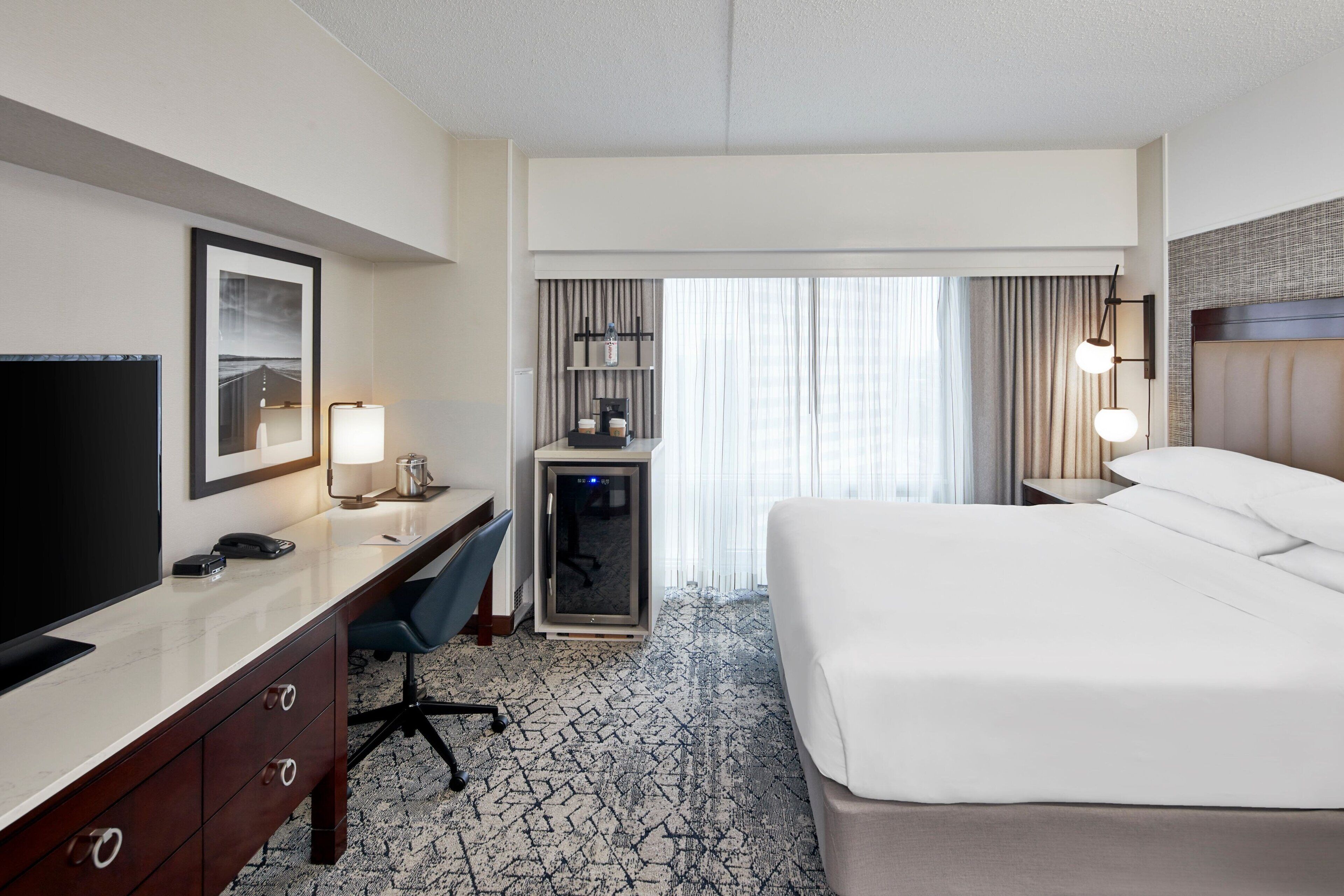 A hotel room with a bed, desk, chair, TV, and a minibar.
