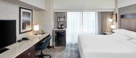 A hotel room with a bed, desk, chair, TV, and a minibar.