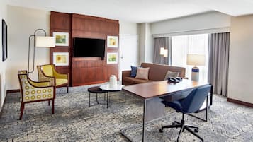 A hotel room with a sofa, a desk, a chair, a TV, and a lamp.