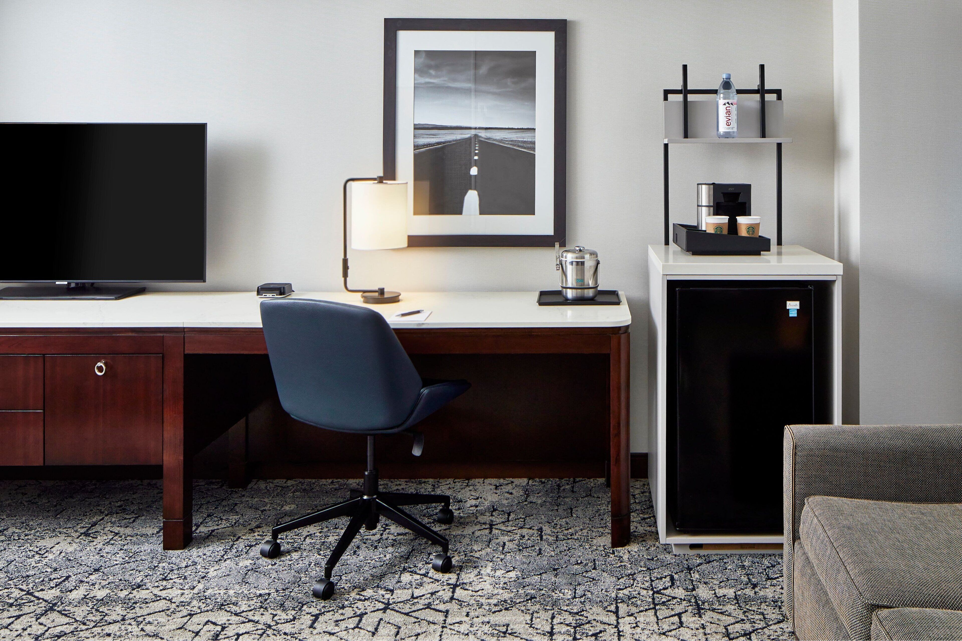 A hotel room with a wooden desk, a black office chair, a flat-screen TV, a framed picture on the wall, a coffee machine, and a mini-fridge.