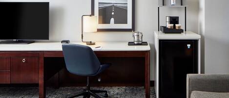 Una habitación de hotel con un escritorio de madera, una silla de oficina negra, un televisor de pantalla plana, un cuadro enmarcado en la pared, una cafetera y un refrigerador pequeño.