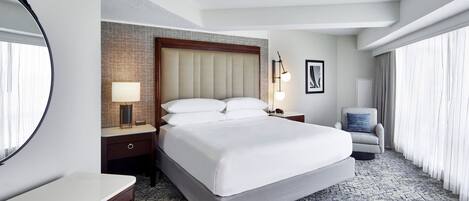 Habitación de hotel con una cama grande, una mesita de noche, un escritorio, una silla, un espejo amplio y una ventana con cortinas.
