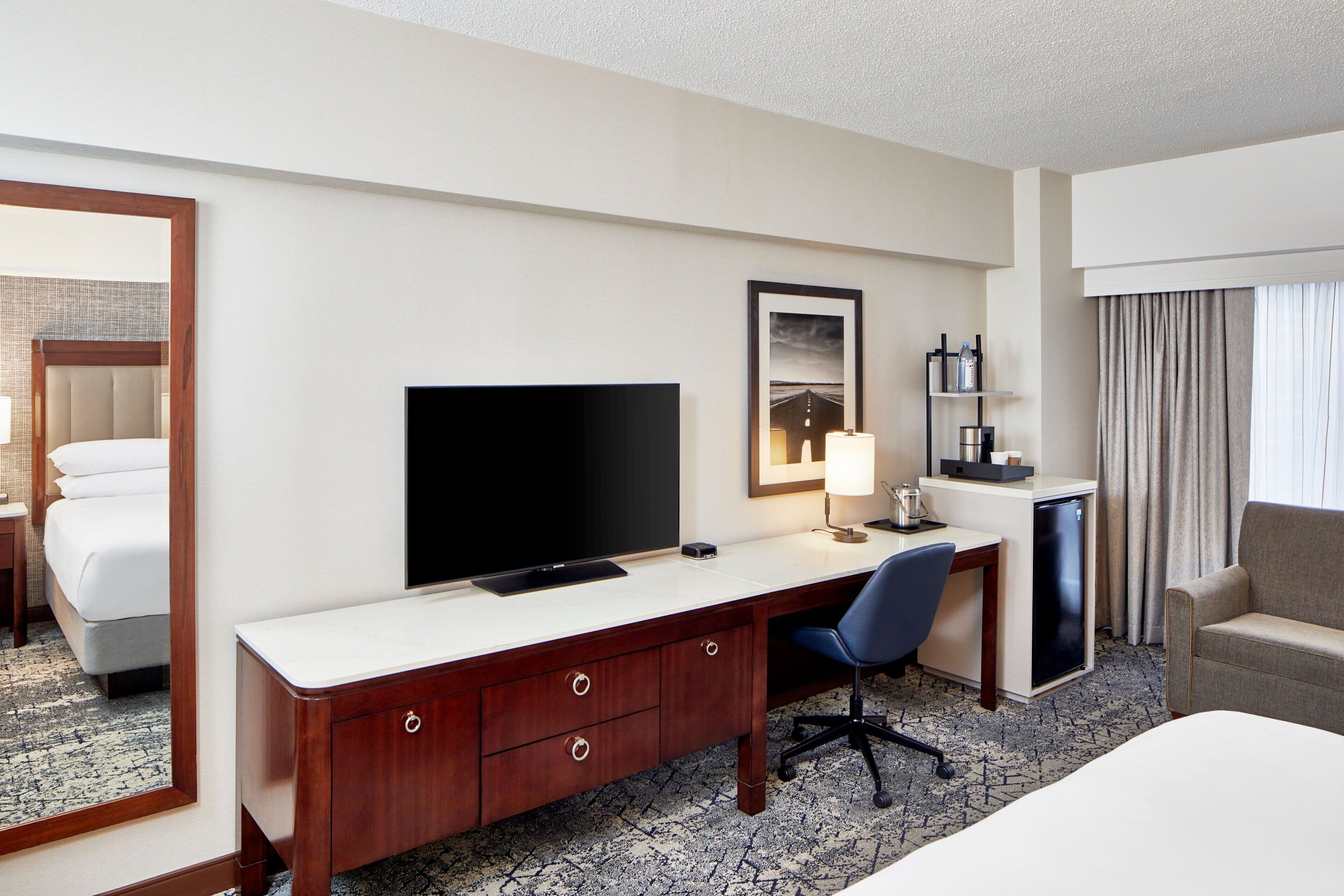 Una habitación de hotel con cama, escritorio y silla, televisión, refrigerador pequeño, espejo y ventana con cortinas.