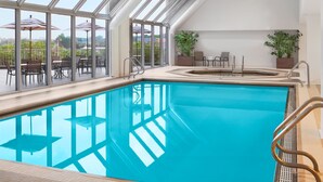 An indoor swimming pool with clear glass walls, a hot tub, and outdoor seating with umbrellas.