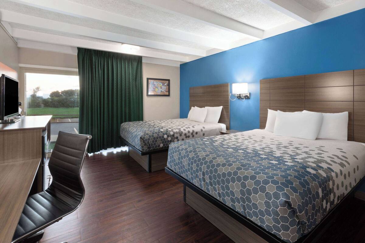 A hotel room with two beds, a desk, a chair, and a large window with a view of a green landscape.