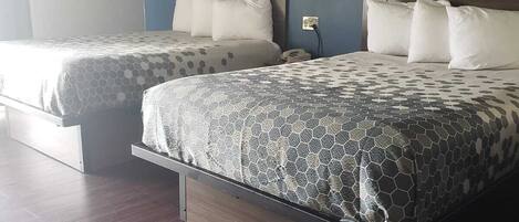 A hotel room with two beds, each with a wooden headboard and a patterned bedspread. There is a nightstand with a lamp between the beds, and a wall-mounted light fixture above the headboard.