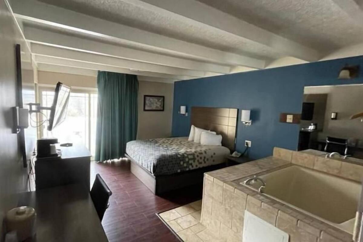 A hotel room with a bed, a desk, a chair, a large window with curtains, a bathroom with a bathtub, and a mirror.