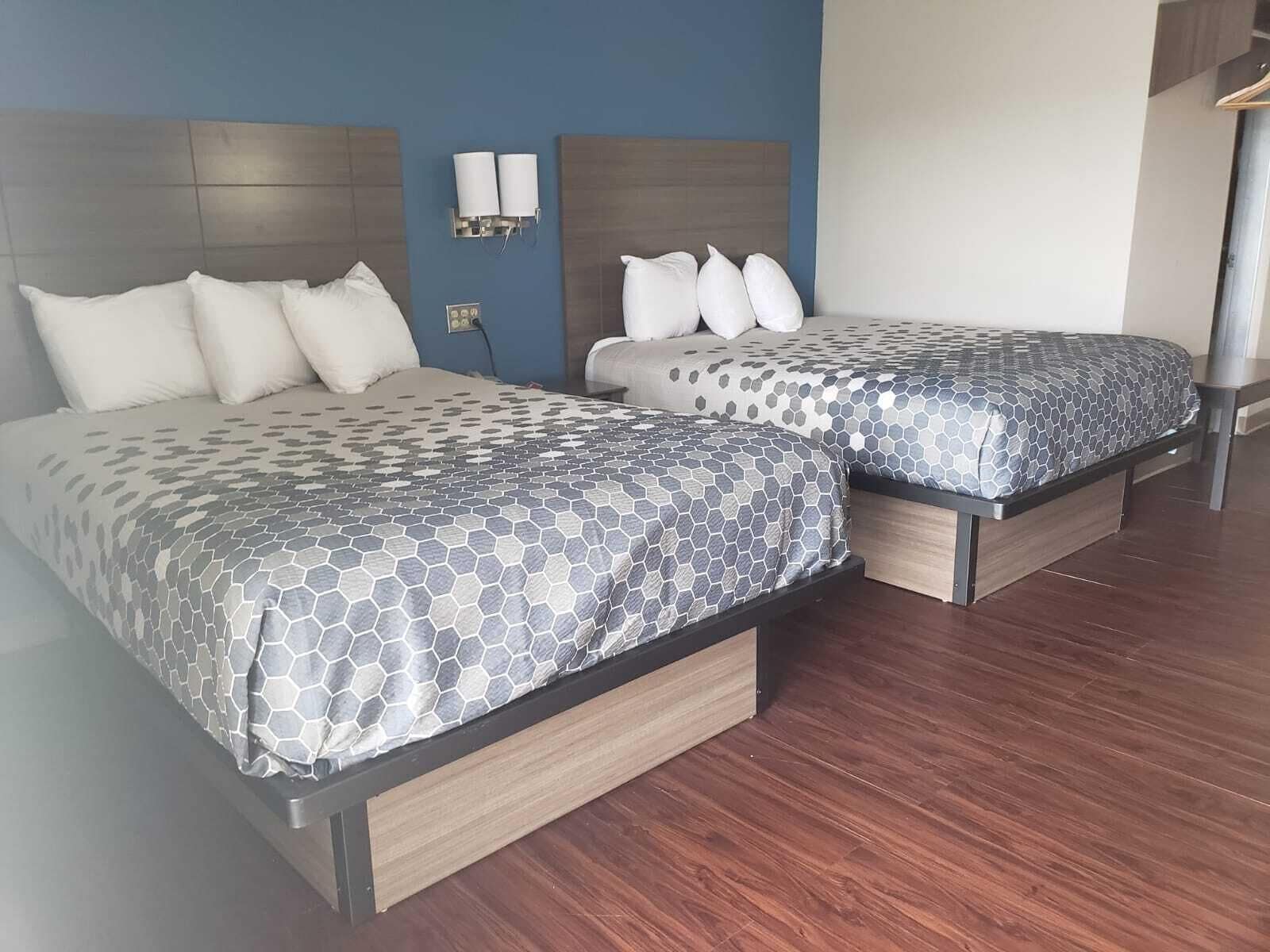 Zwei Betten mit grau-weiß gemusterten Bettbezügen, Holz-Kopfteilen und einer blauen Wand mit Lichtschalter.