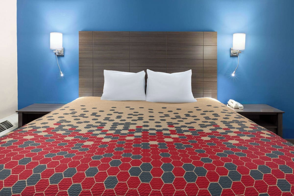 A hotel room with a large bed, two white pillows, a patterned red and gray bedspread, a wooden headboard, and two bedside lamps.