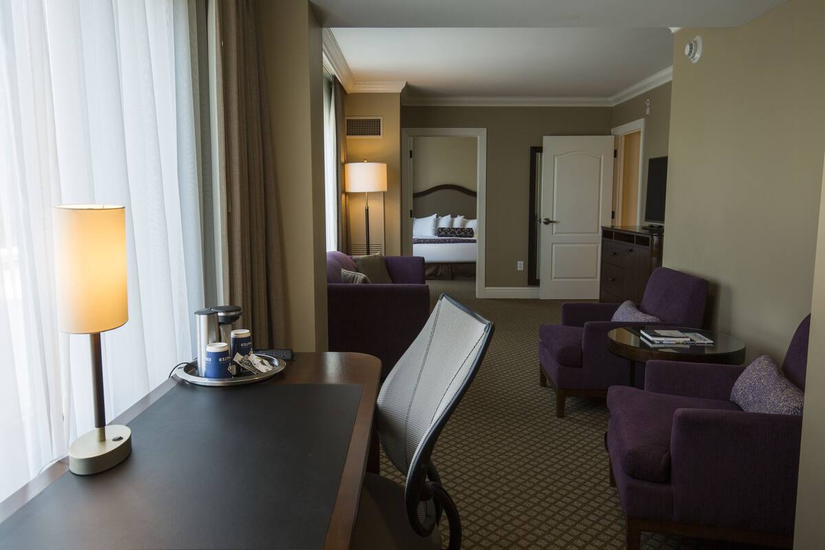A hotel room with a bed, two purple armchairs, a small table, a lamp, and a view of the hallway.
