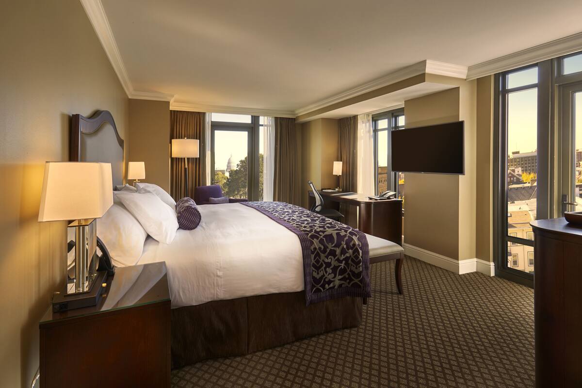 A hotel room with a large bed, a desk, a TV, and a view of the city.