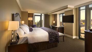 A hotel room with a large bed, a desk, a TV, and a view of the city.