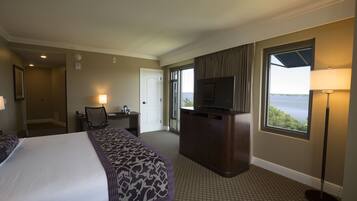 A hotel room with a large bed, a desk with a chair, a television, and a window with a view of the water.