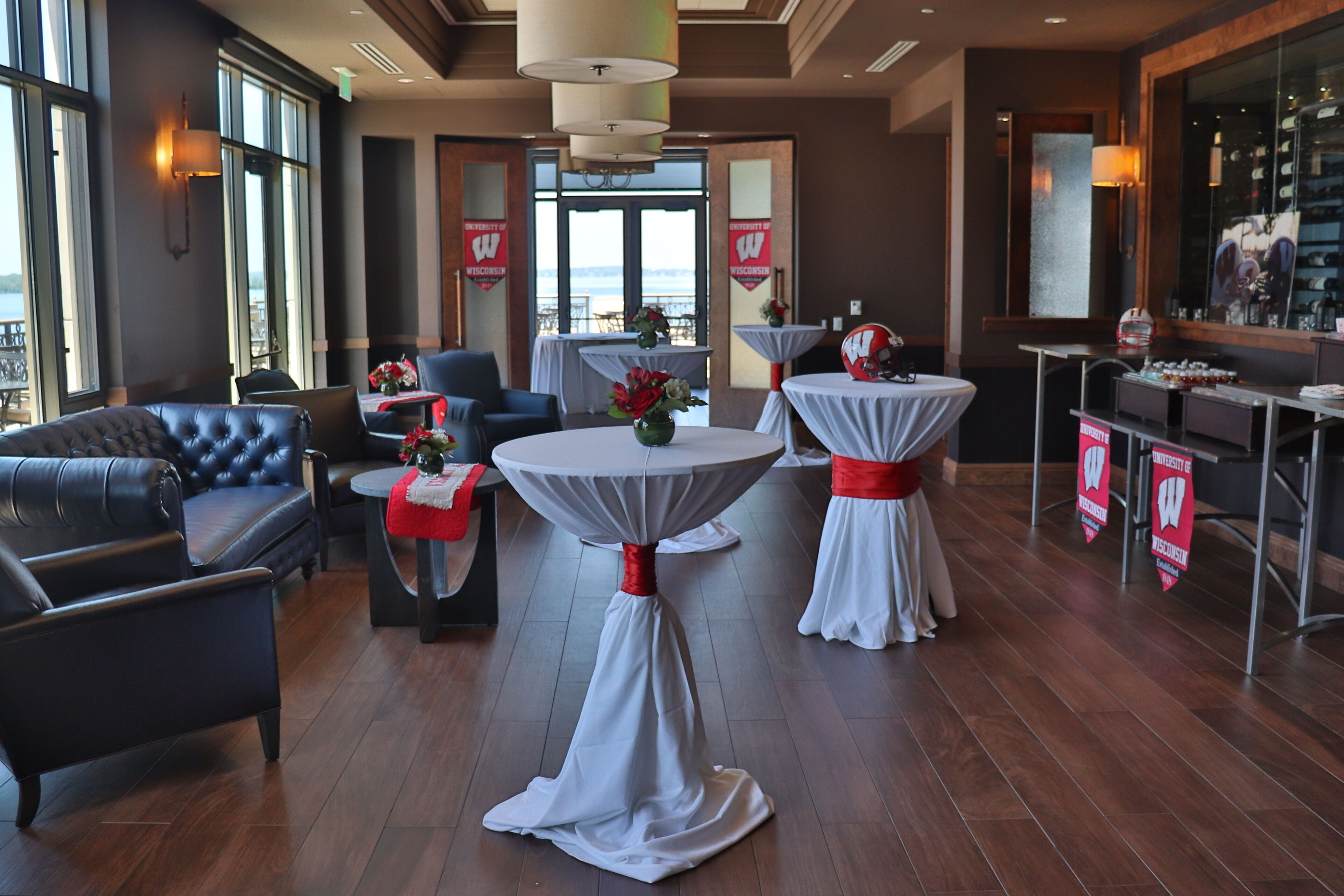 Un lobby de hotel moderno con asientos de cuero, mesas redondas con manteles blancos y fajas rojas, y grandes ventanales que ofrecen vistas al exterior.