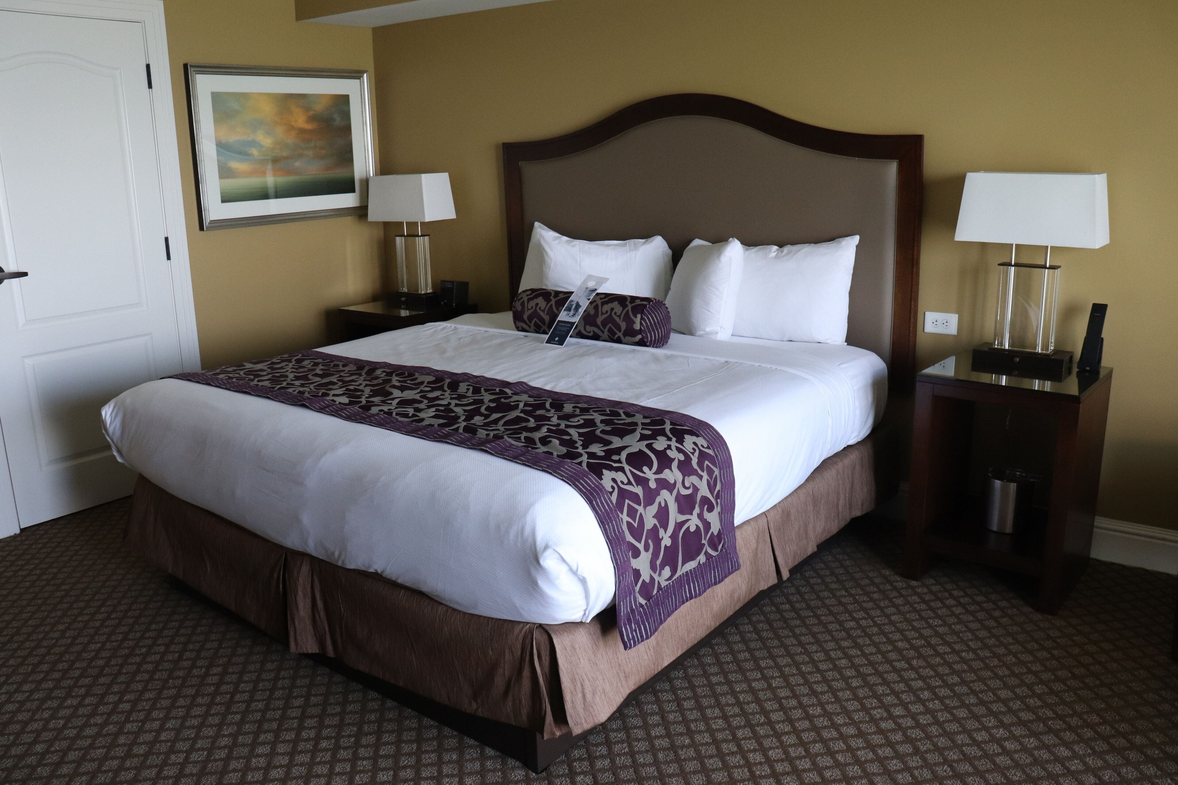 A hotel room with a large bed, two bedside tables with lamps, a framed picture on the wall, and a patterned carpet.